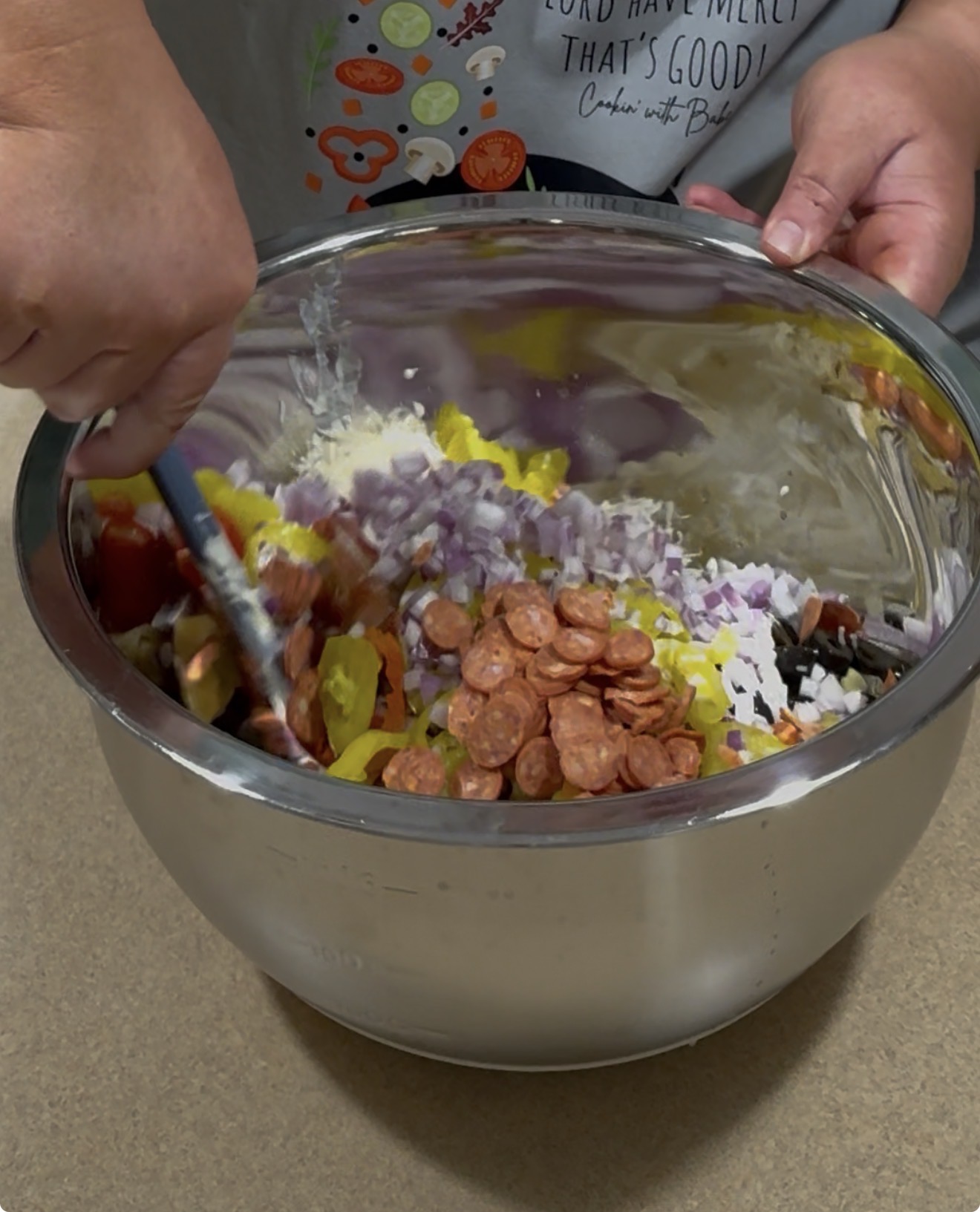 A person stirs a mixture of chopped onions, mini pepperoni slices, and yellow peppers in a large metal bowl on a kitchen counter.