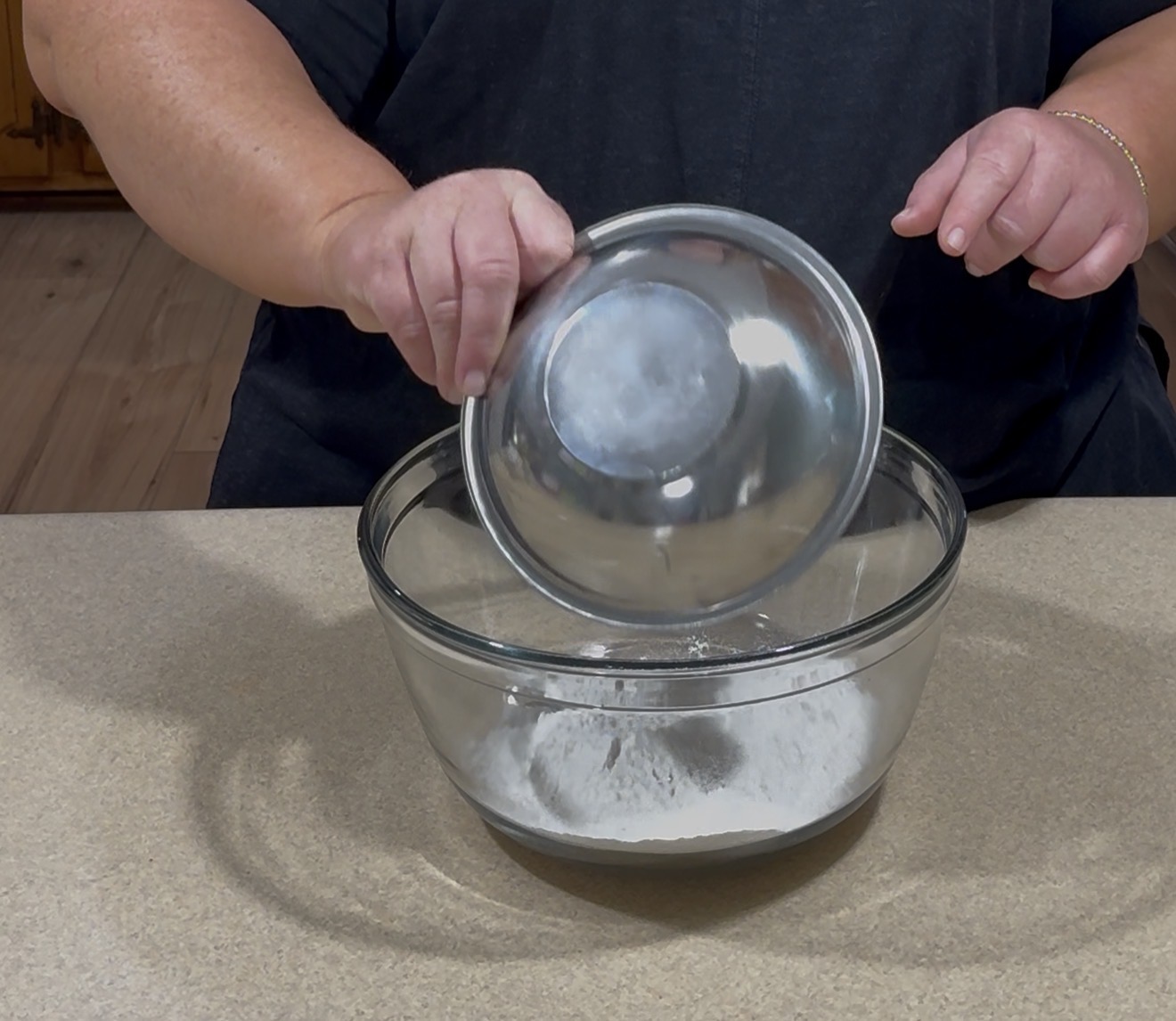 A person pours flour from a metal bowl into a glass mixing bowl on a kitchen counter.