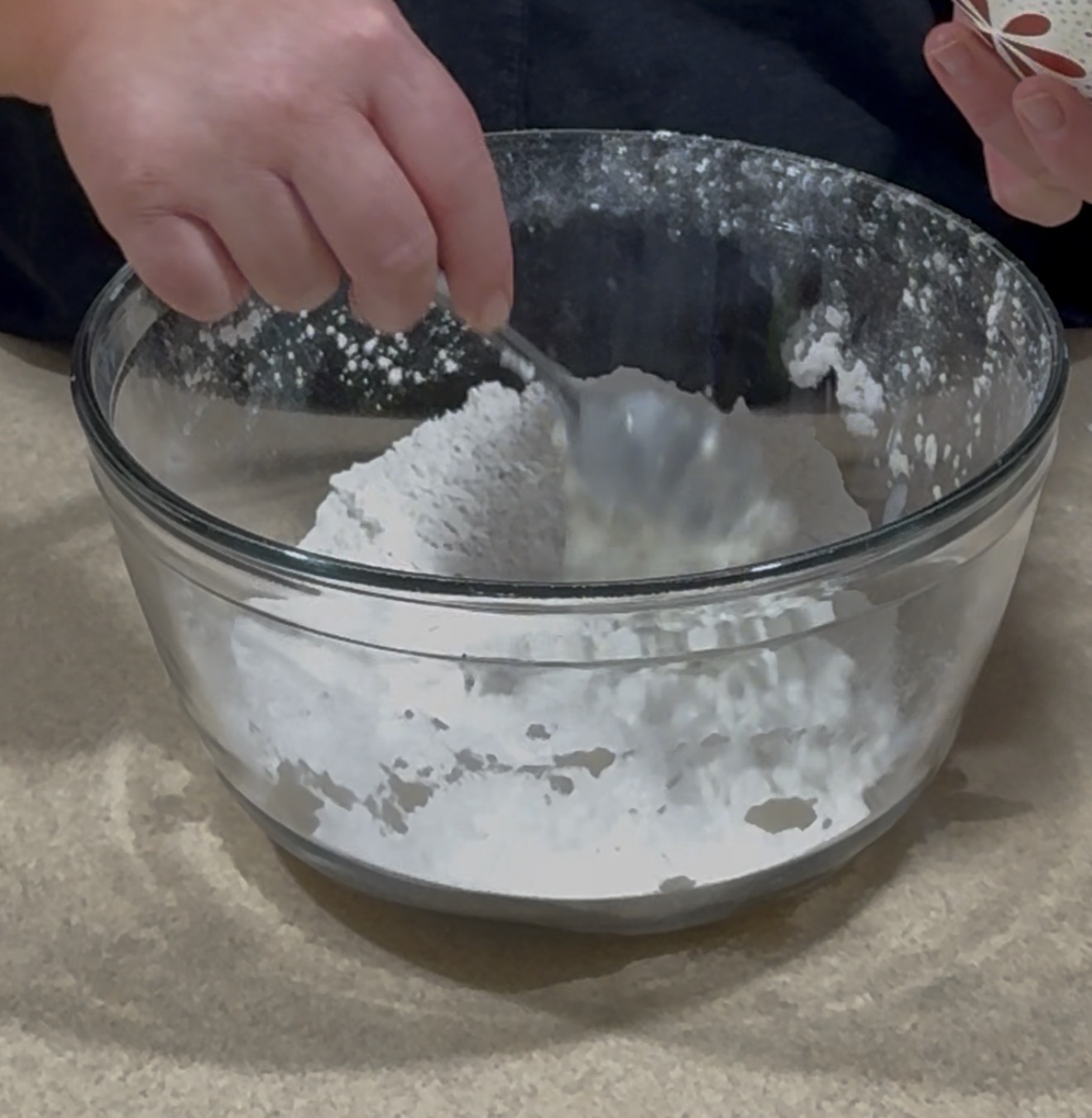 A person stirs white flour in a clear glass mixing bowl with a spoon on a beige countertop. Another hand holds a small container near the bowl.