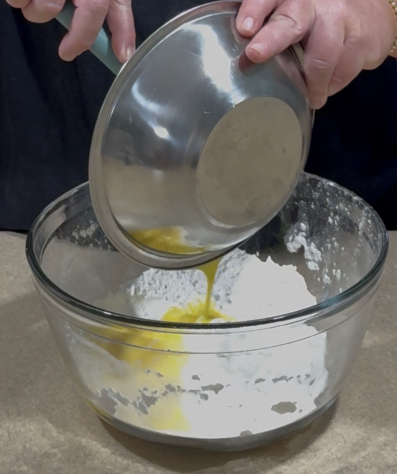 A person pours a bowl of beaten eggs into a larger glass bowl containing flour, preparing to mix ingredients for baking.
