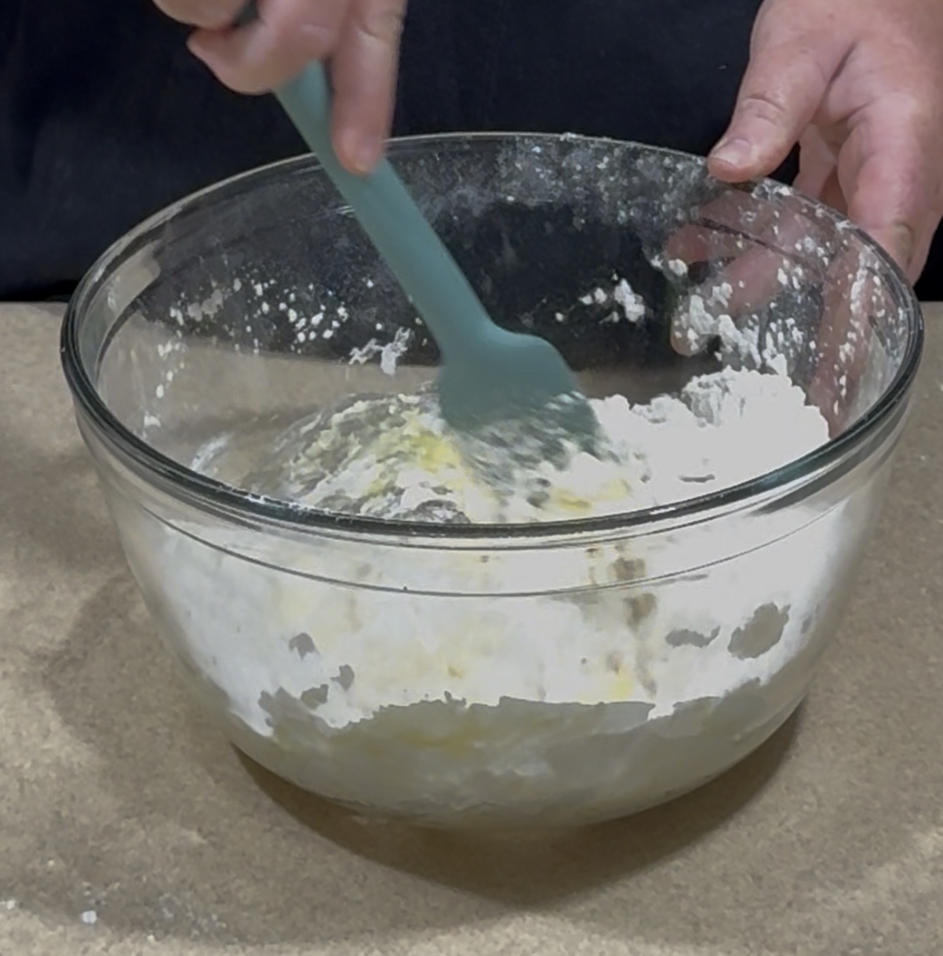 A person uses a teal spatula to mix flour and other ingredients in a large glass bowl placed on a countertop.