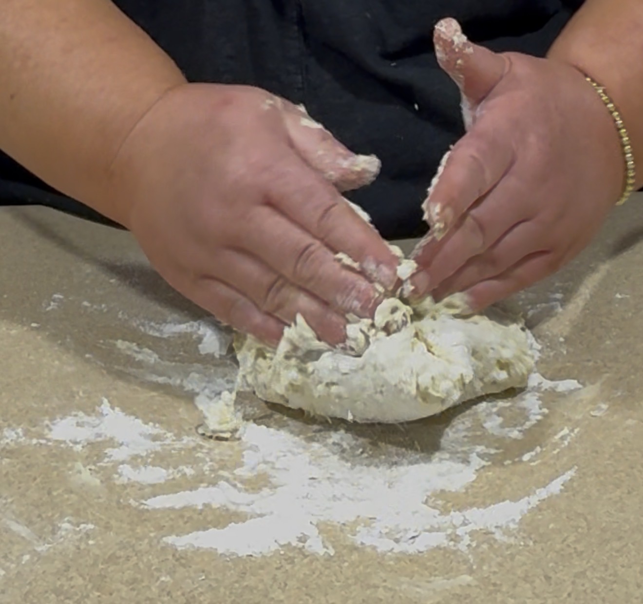Hands kneading a ball of dough on a floured countertop, with bits of flour scattered around and some dough sticking to the fingers.