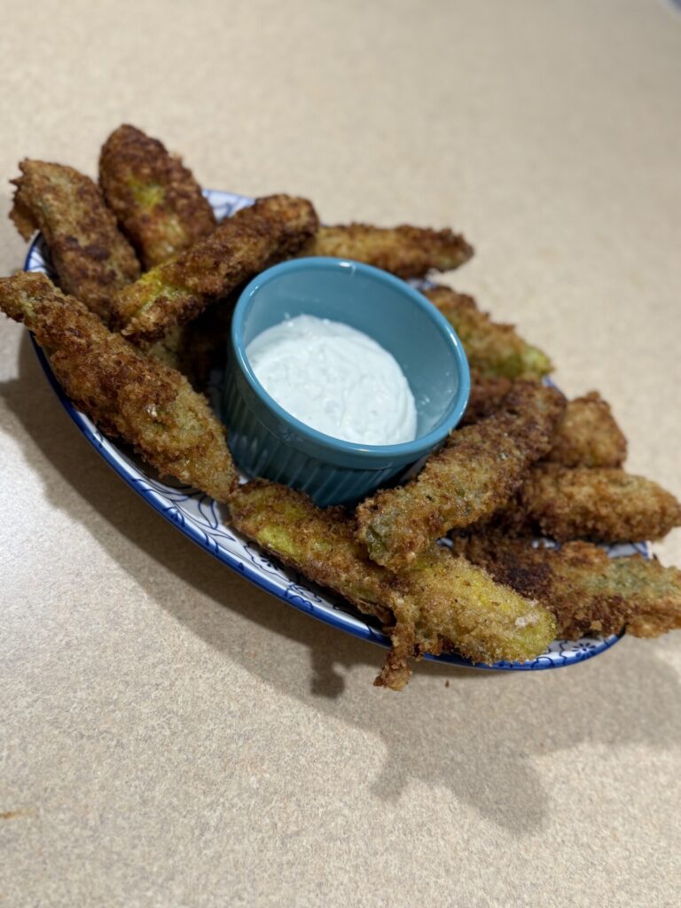 A plate of golden-brown fried pickles is arranged around a small blue bowl filled with creamy dipping sauce, set on a beige countertop.