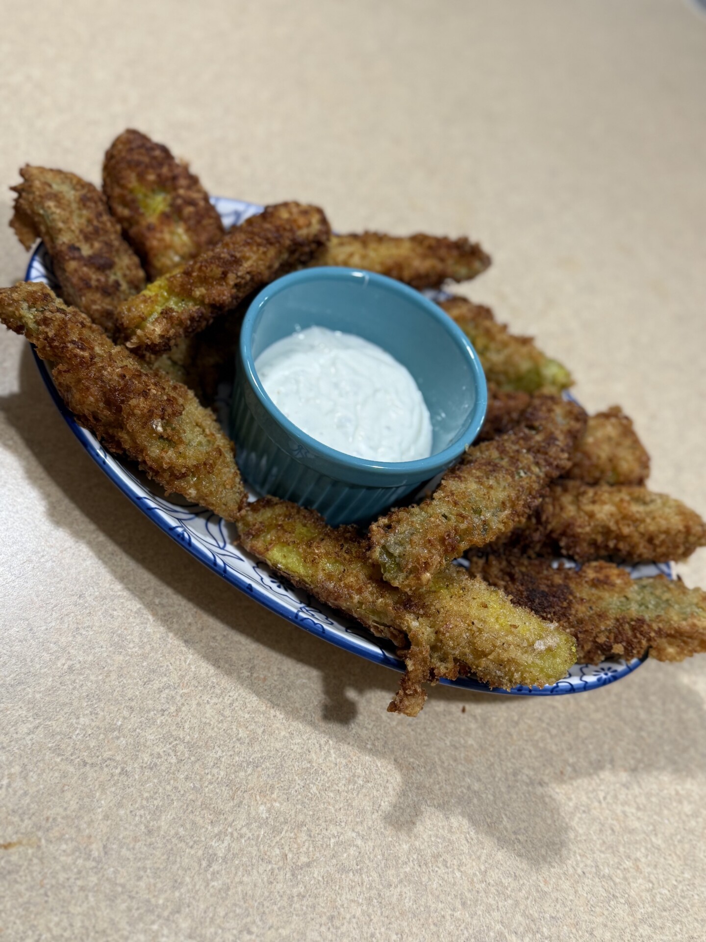 A plate of golden-brown fried pickles is arranged around a small blue bowl filled with creamy dipping sauce, set on a beige countertop.
