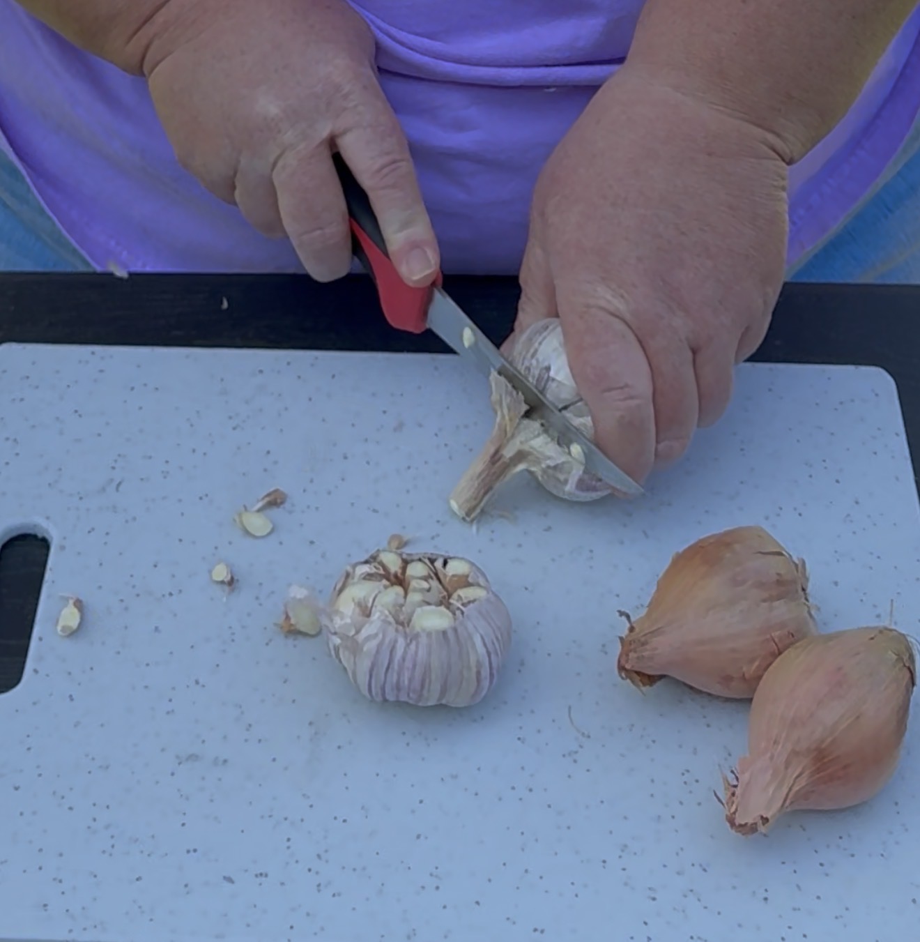 A person in a purple shirt slices the top off a head of garlic on a white cutting board, with two shallots beside the garlic.
