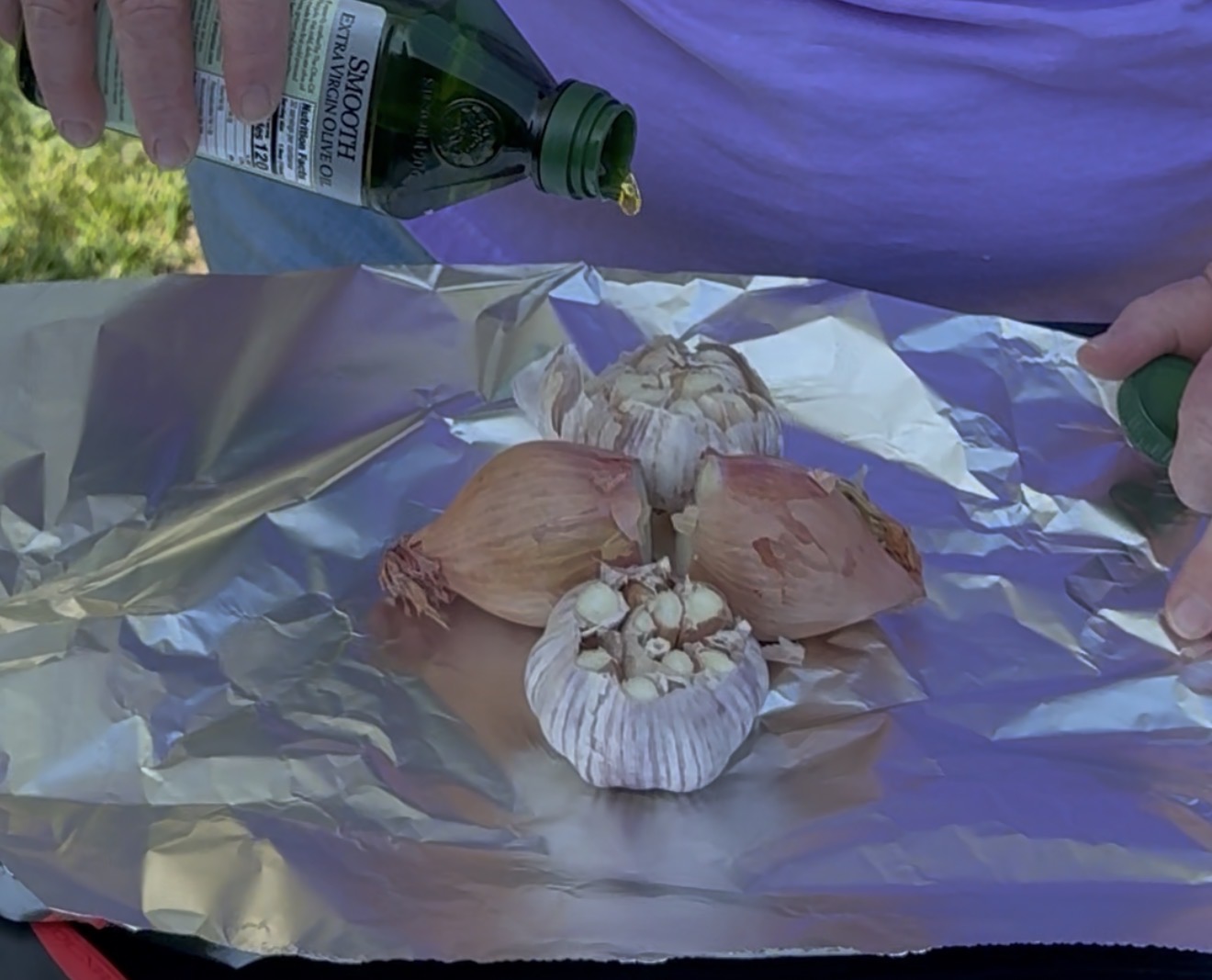 A person pours olive oil over heads of garlic and shallots placed on a sheet of aluminum foil, preparing them for roasting.