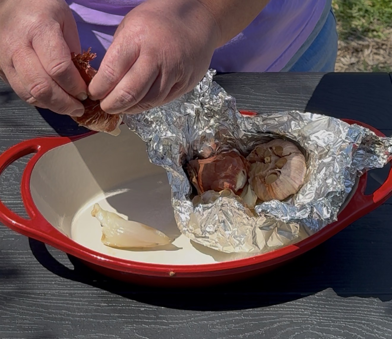 A person peels roasted garlic from foil over a red baking dish on a black outdoor table. One bulb is exposed, with garlic cloves and wrappers visible inside the foil.