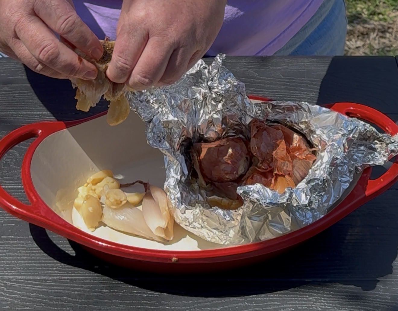 A person peels roasted garlic cloves from a foil packet into a red, oval baking dish; other peeled garlic cloves are already in the dish, set on a black table outdoors.