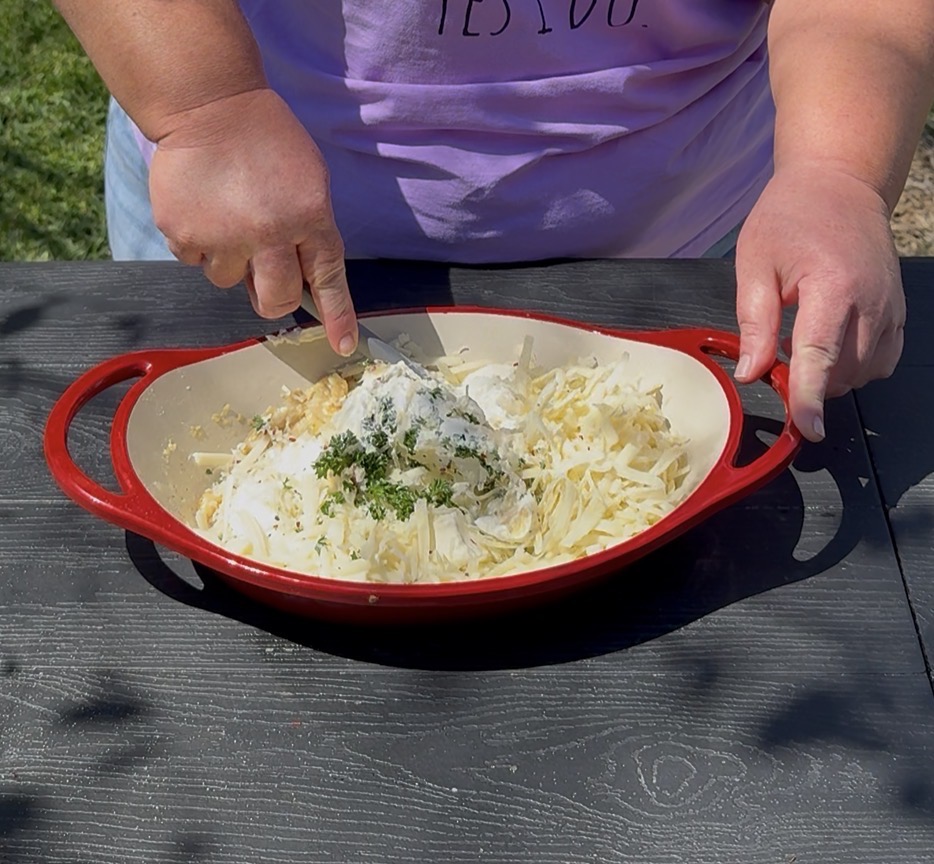 A person mixes shredded potatoes, herbs, and a creamy sauce in a red baking dish on a black table outdoors. Only their hands and part of a purple shirt are visible.