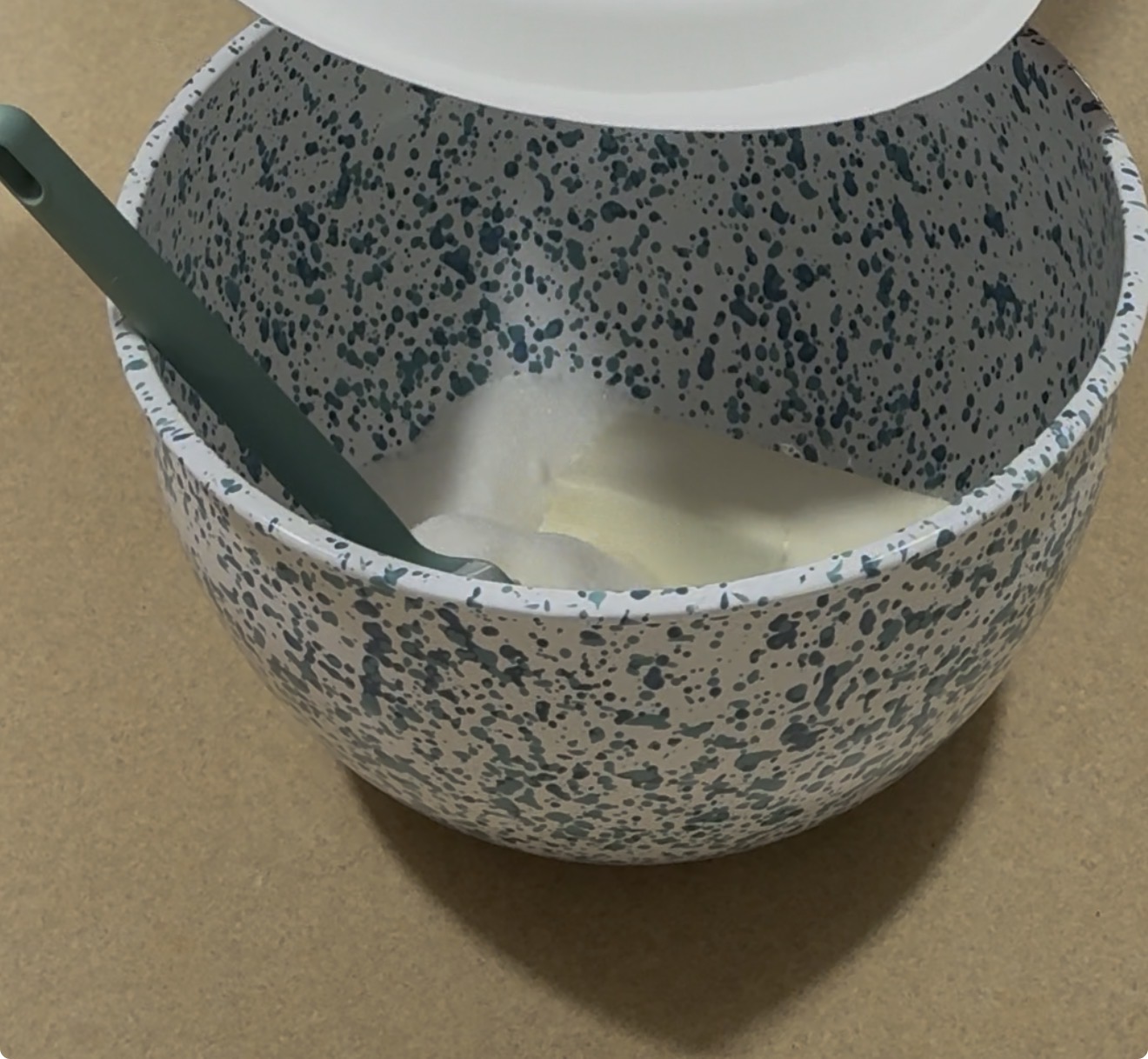 A speckled mixing bowl with a green spatula rests on a tan surface. The bowl contains a mound of white sugar and a piece of butter. The lid is partially covering the bowl.