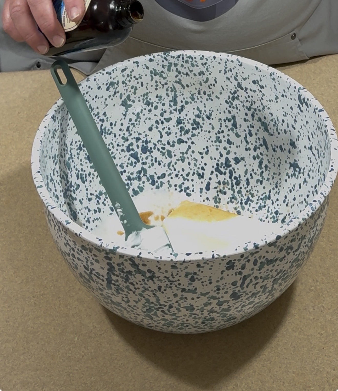 A person pours vanilla extract from a bottle into a large white and blue speckled mixing bowl containing white ingredients, using a green spatula to mix.