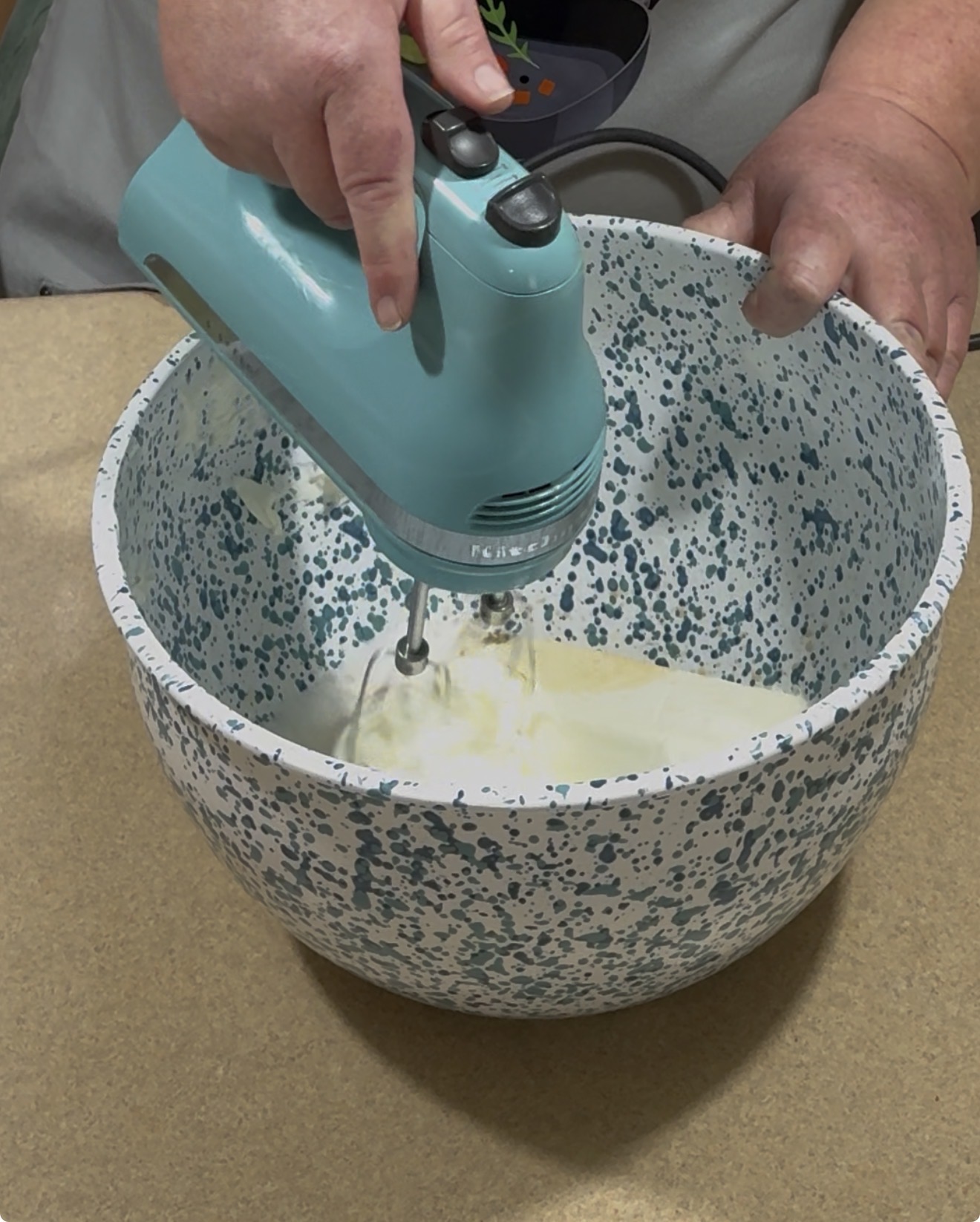 A person uses a teal electric hand mixer to blend ingredients in a large white and blue speckled mixing bowl on a beige countertop.