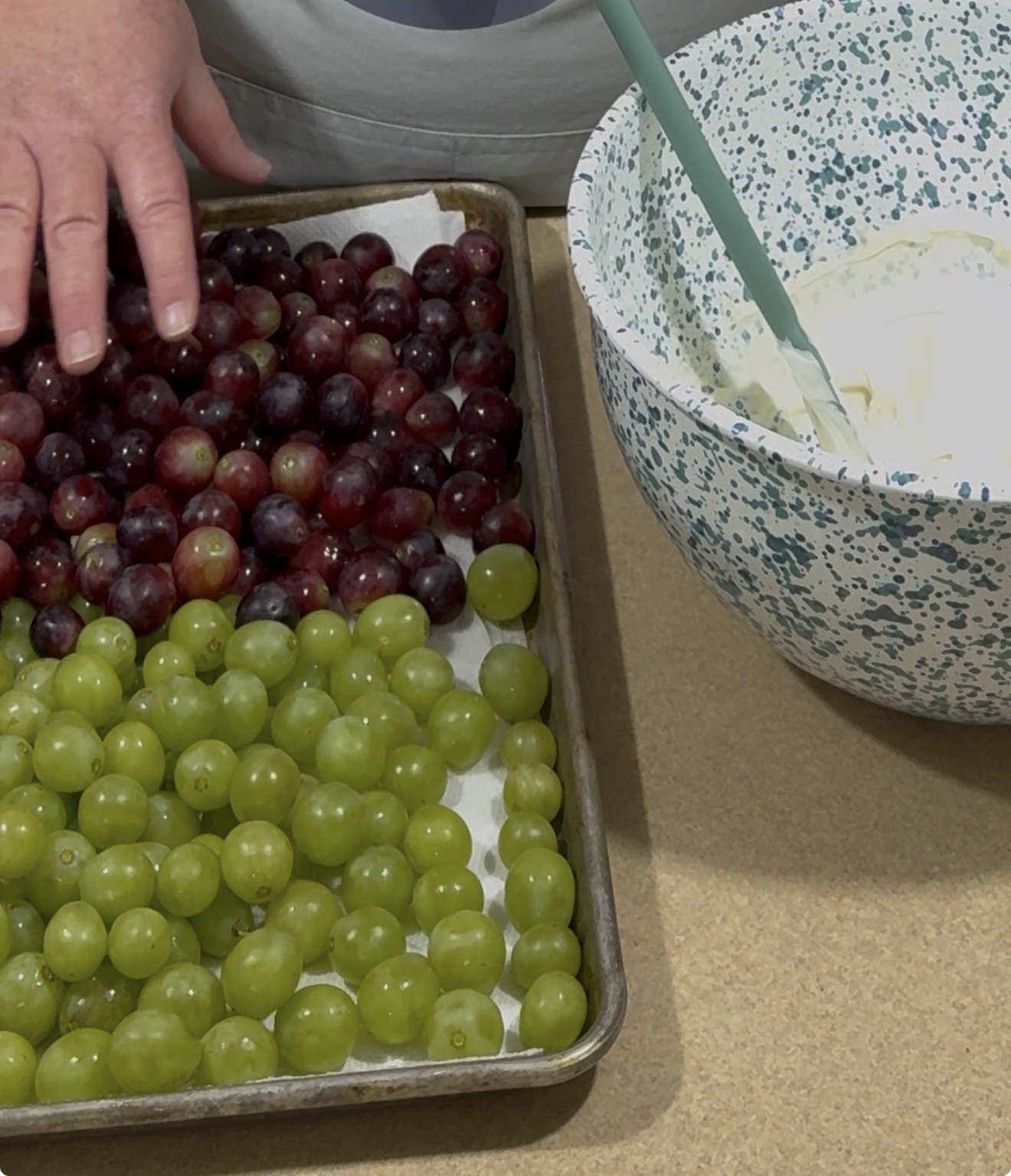 A hand arranges red and green grapes on a baking sheet lined with parchment paper. Next to the tray is a bowl filled with a white mixture and a mixing spoon.