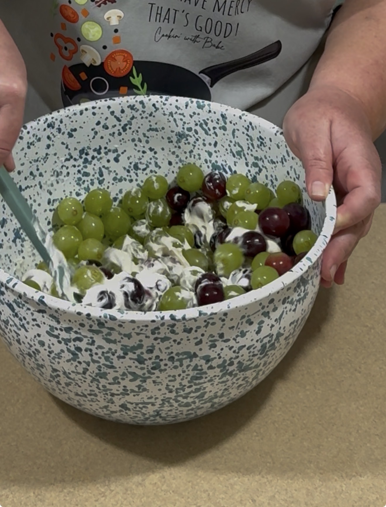 A person stirs green and red grapes with a creamy mixture in a speckled mixing bowl. The person is wearing a gray shirt with colorful kitchen graphics and text.