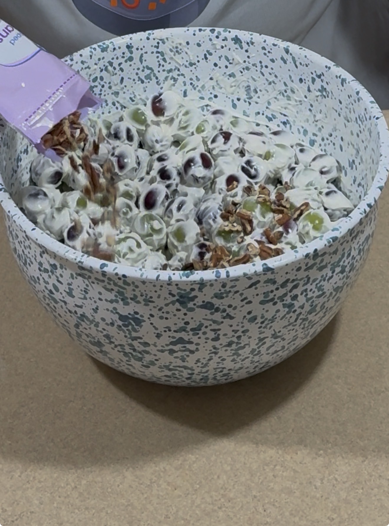 A person adds chopped pecans from a packet into a large bowl of creamy grape salad on a countertop.