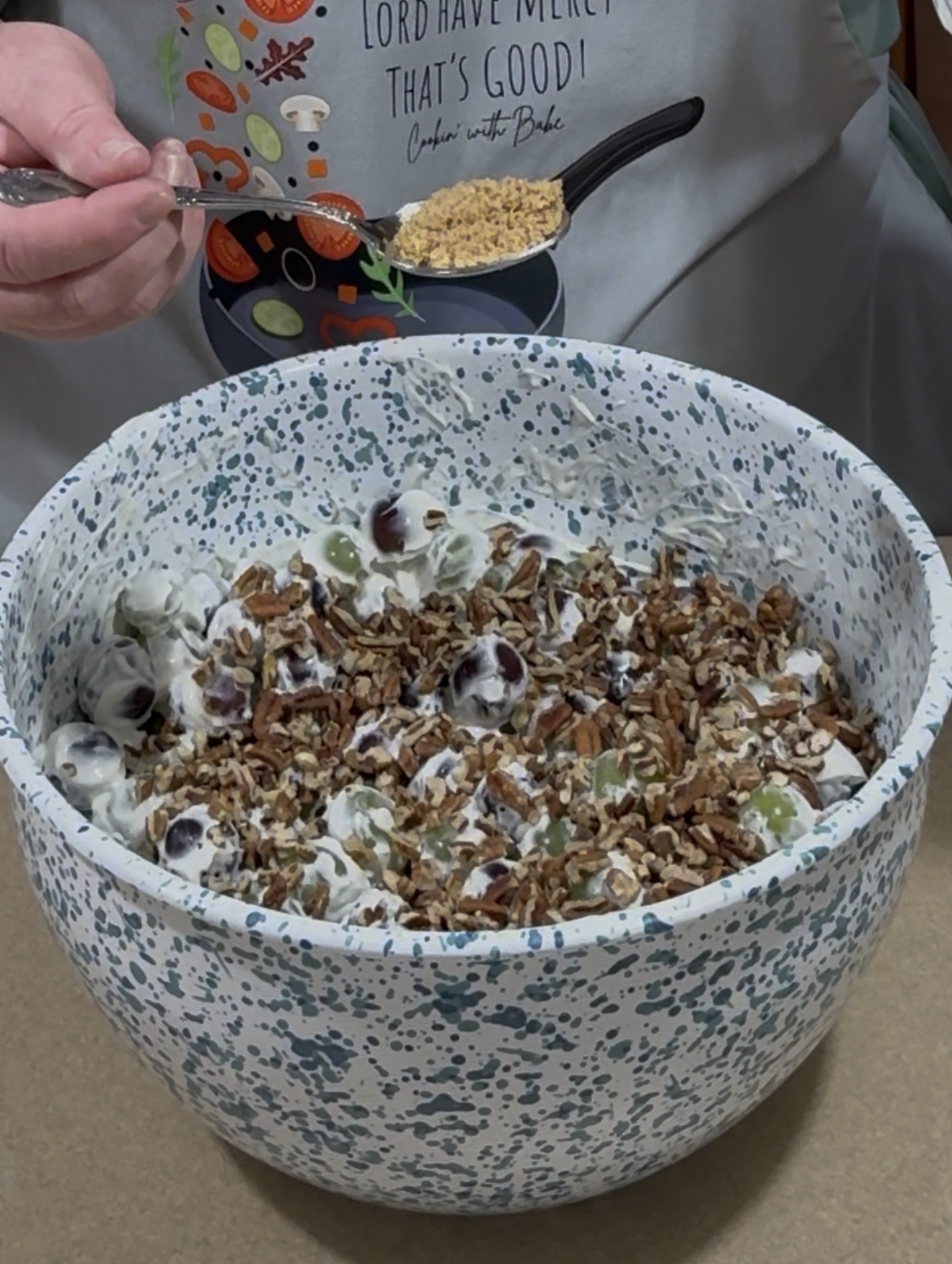 A person wearing an apron holds a spoon above a large speckled bowl filled with a creamy salad topped with chopped pecans and grapes.