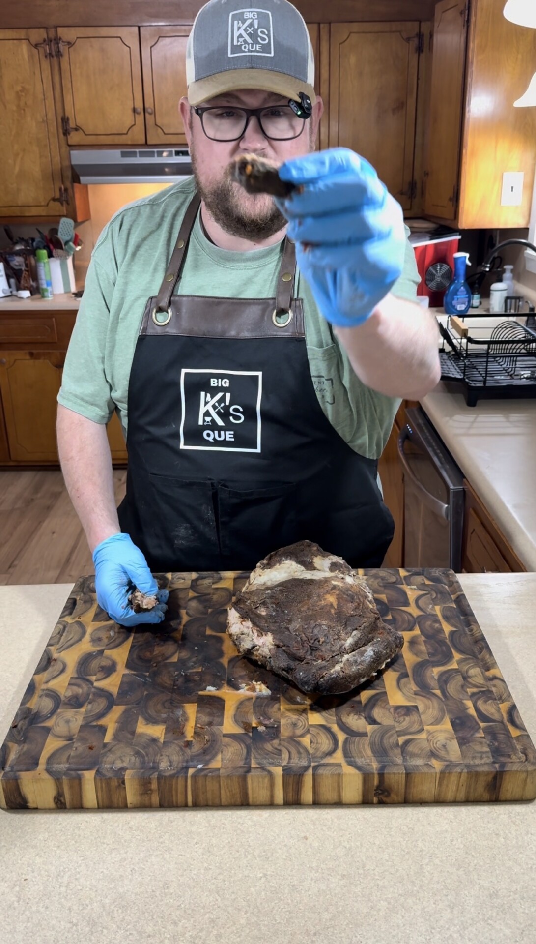 A man in a green shirt, black apron, and blue gloves holds a piece of cooked meat toward the camera. A large cooked meat rests on a wooden cutting board in a kitchen setting.