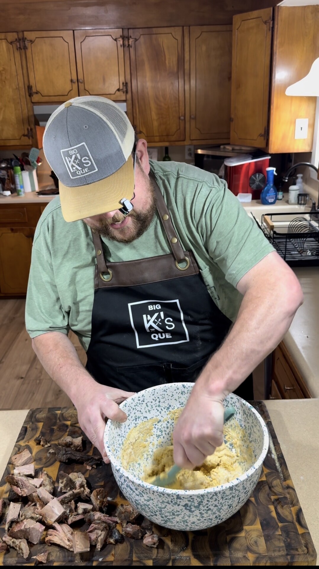 A man in a green shirt, black apron, and a gray hat stirs a large bowl of food in a kitchen. Chopped meat is on a cutting board in front of him, and wooden cabinets and kitchen items are in the background.