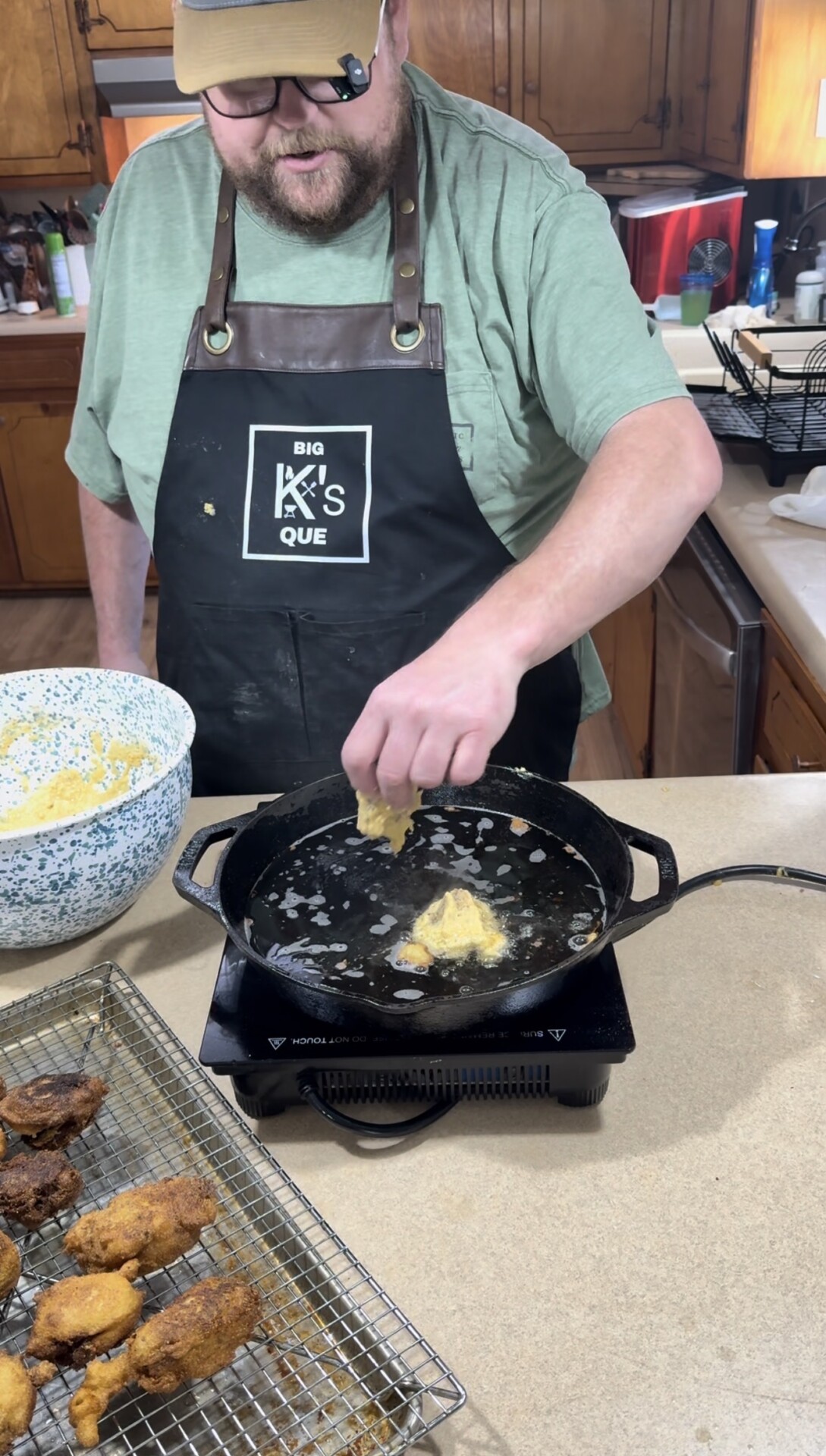 A person wearing a green shirt and a black “Big K’s Que” apron fries battered food in a black skillet on a stovetop, with a bowl of batter and a cooling rack of cooked food nearby in a kitchen.