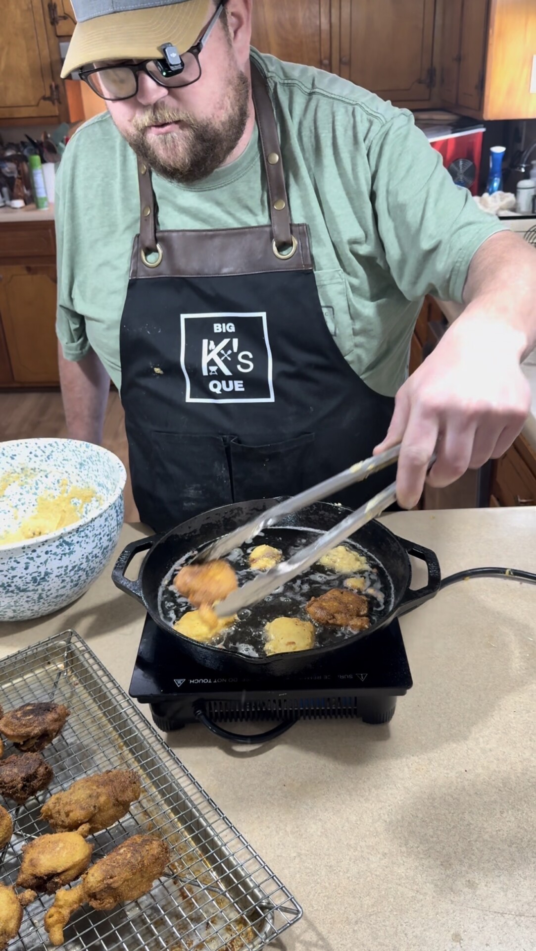 A person wearing a green shirt and black apron is frying battered food in a black pan using tongs. There is a bowl of batter and a cooling rack with fried pieces on a kitchen counter.