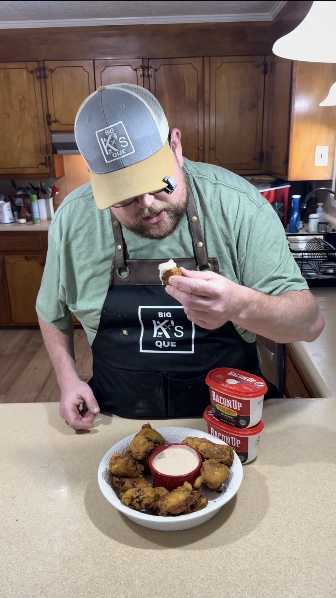 A man in a green shirt and apron labeled “Big K’s Que” holds a fried food item over a bowl of dipping sauce, standing in a kitchen with tubs of Bacon Up on the counter.