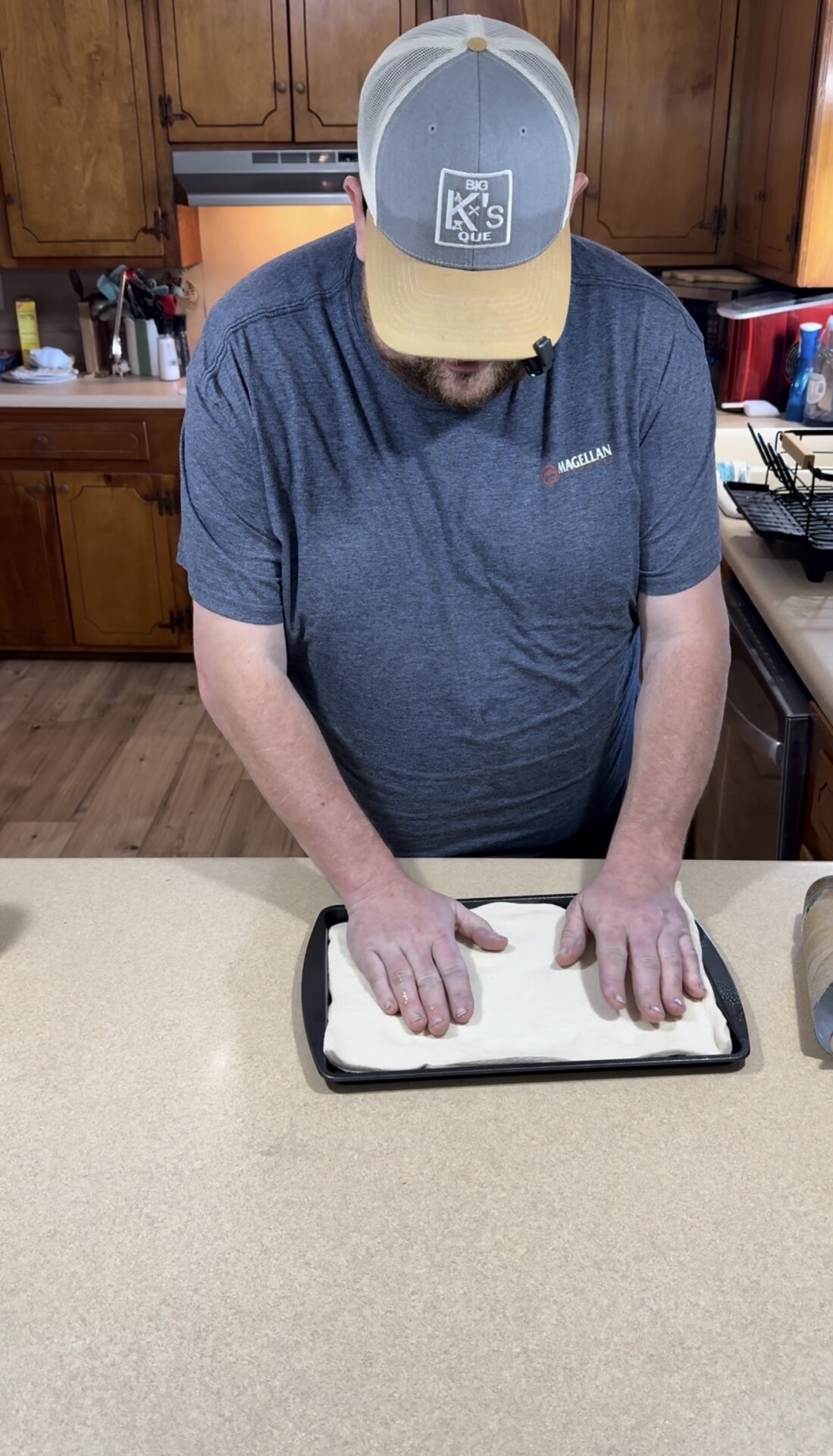 A person wearing a gray shirt and hat presses dough flat into a rectangular baking pan on a kitchen counter. The kitchen has wooden cabinets and various utensils in the background.