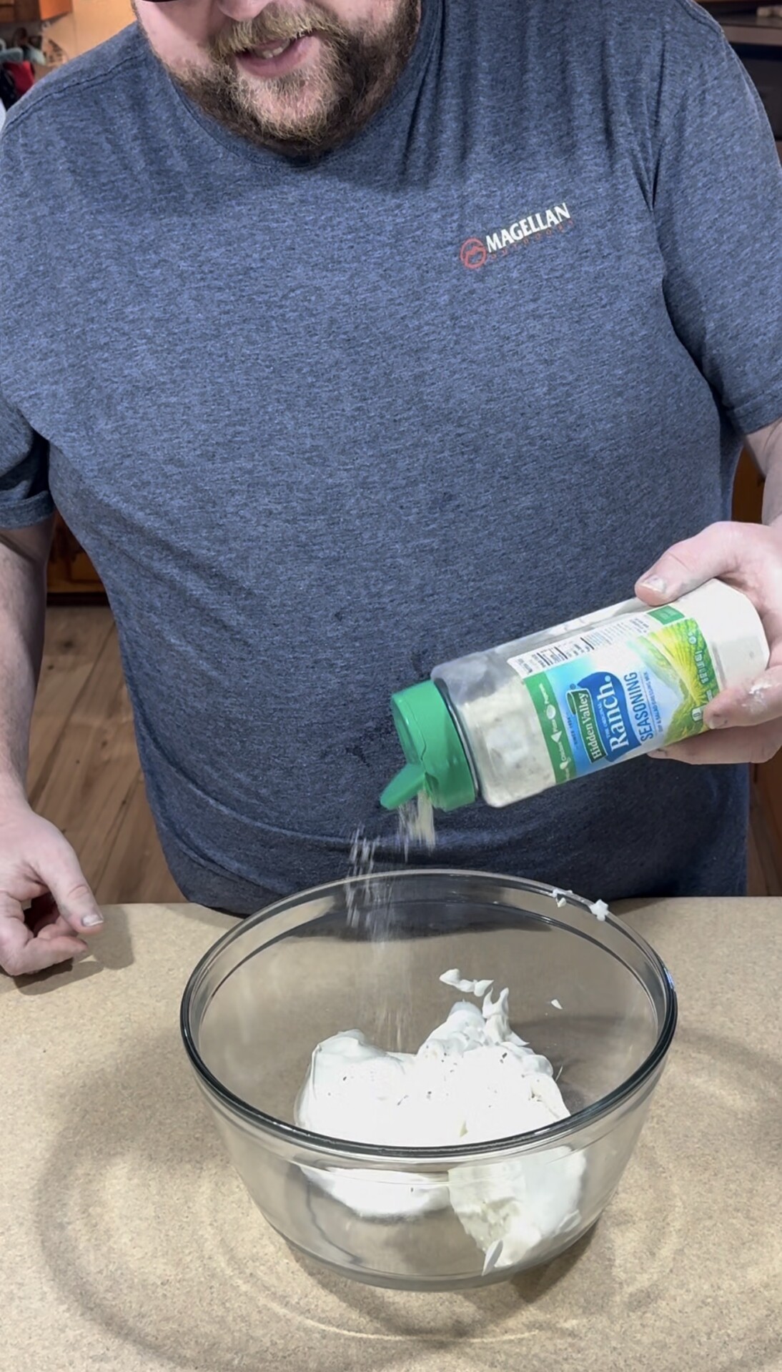 A person sprinkles ranch seasoning from a plastic container into a glass bowl filled with white cream or dip on a kitchen counter.