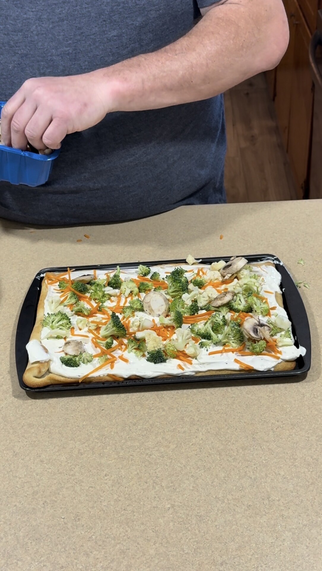 A person in a gray shirt is preparing a rectangular veggie pizza on a baking tray, topping it with fresh vegetables like broccoli, carrots, and mushrooms on a creamy base.