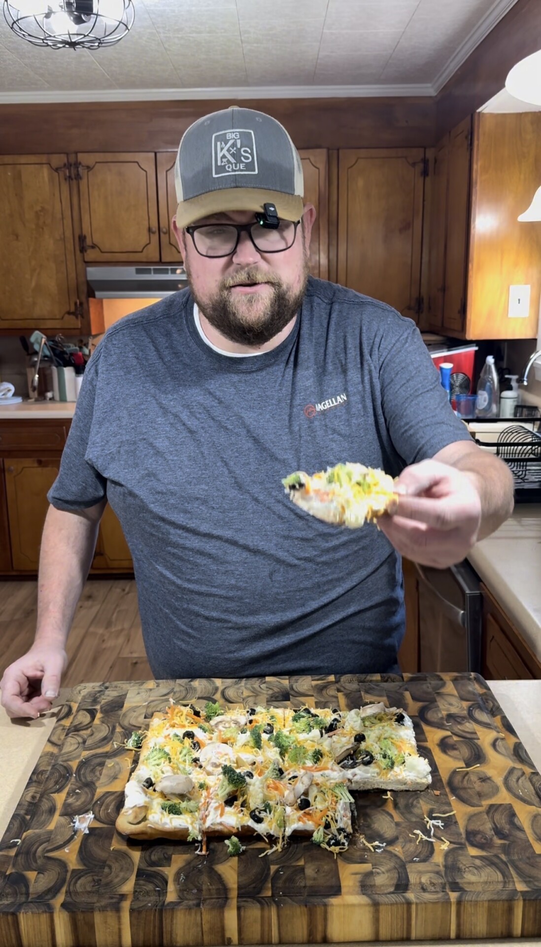 A bearded man wearing glasses, a gray cap, and a gray shirt stands in a kitchen, holding up a piece of loaded pizza or casserole on a spatula, with a cutting board of the dish in front of him.