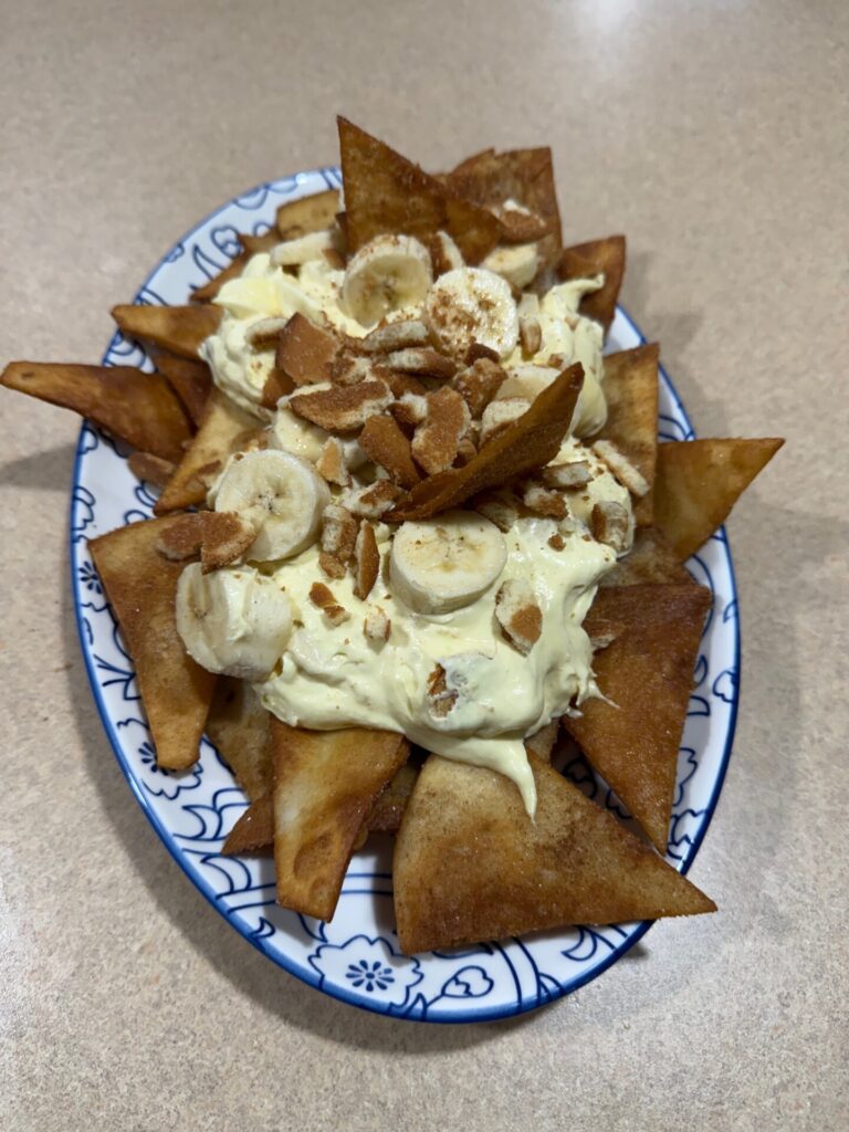 A plate of fried tortilla chips topped with banana slices, creamy pudding, and crushed nuts, served on a decorative oval dish atop a beige countertop.