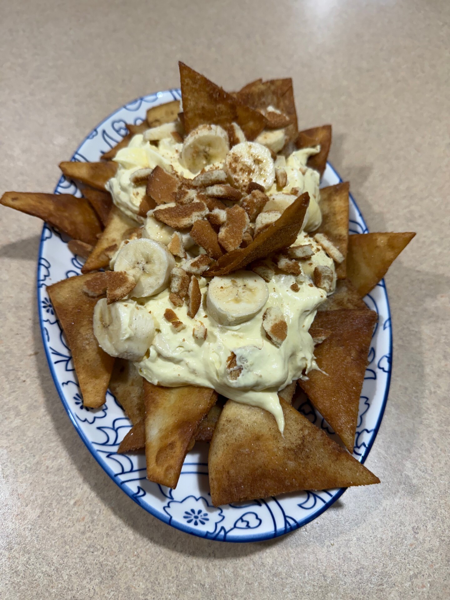 A plate of fried tortilla chips topped with banana slices, creamy pudding, and crushed nuts, served on a decorative oval dish atop a beige countertop.