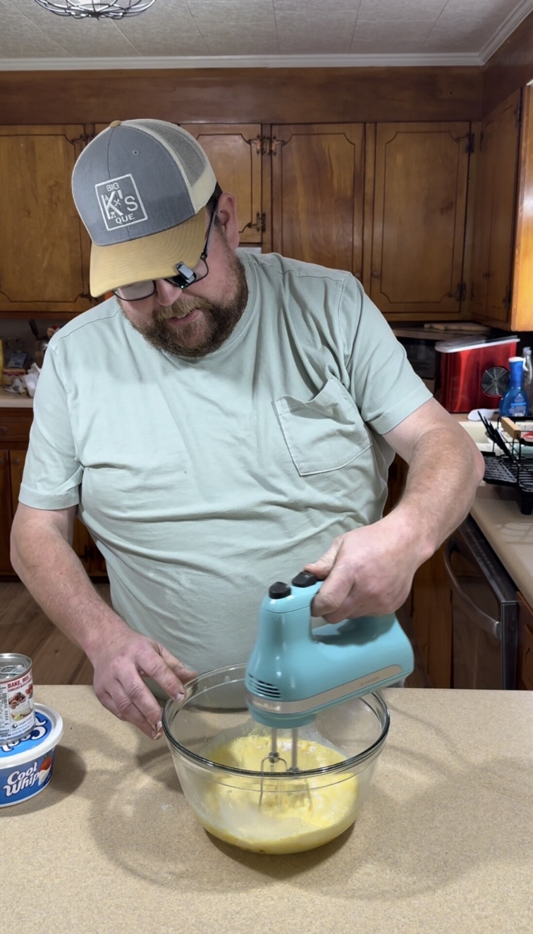 A man in a light green shirt and hat is mixing ingredients in a glass bowl with a blue hand mixer in a kitchen. Various items, including a Cool Whip container, are on the counter nearby.