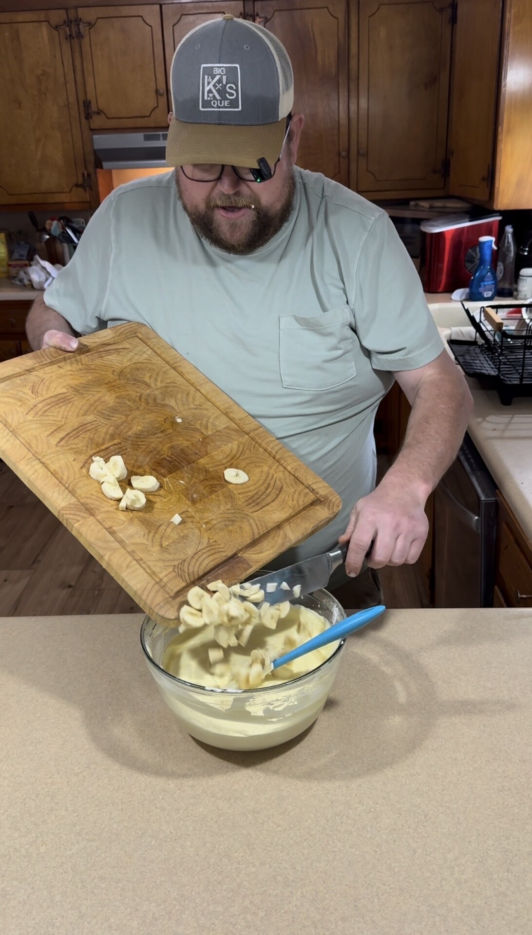 A man in a light green shirt and cap is adding sliced bananas from a wooden cutting board into a glass bowl of batter on a kitchen counter. A blue spatula rests in the bowl.