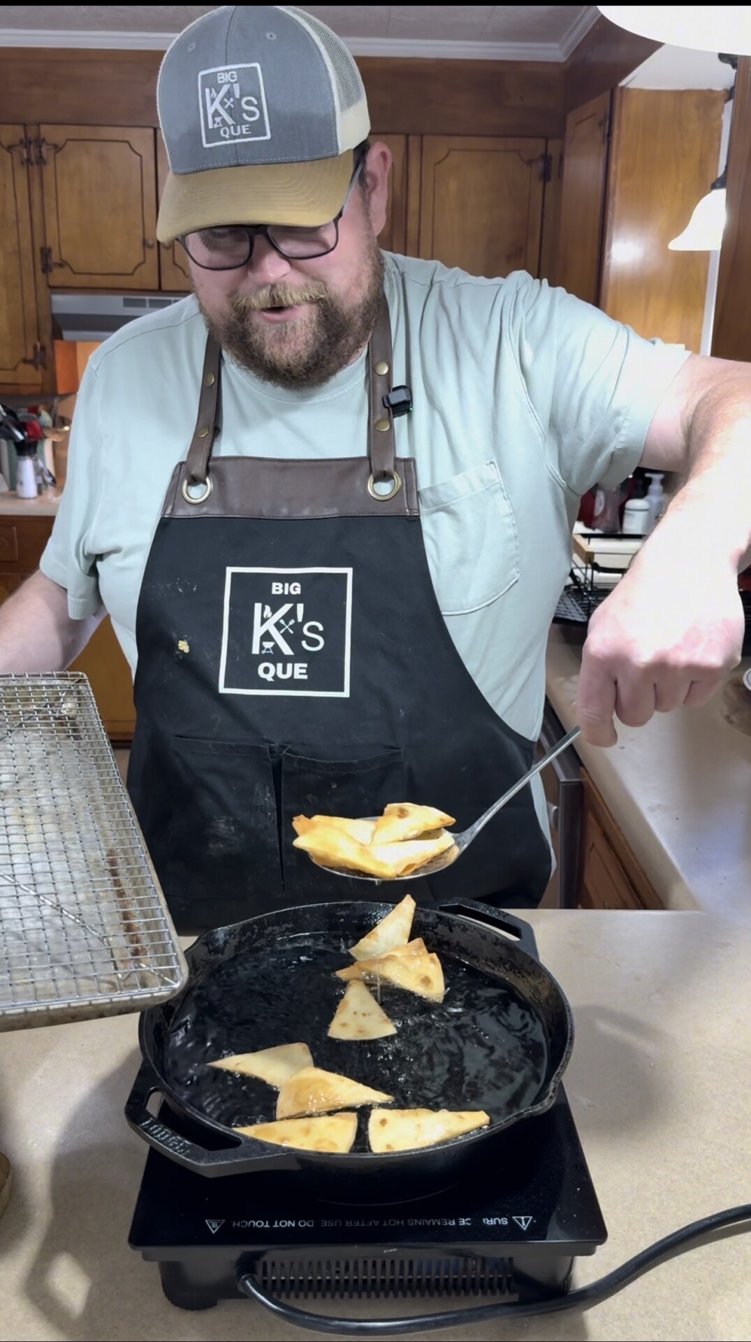 A man wearing a Big Ks Que apron and hat fries tortilla chips in a cast iron skillet, holding a slotted spoon and a cooling rack in a kitchen.