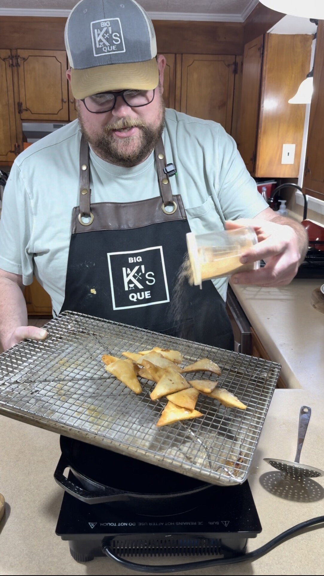 A man wearing glasses, a cap, and a black apron labeled Big Ks Que sprinkles seasoning onto crispy chips on a cooling rack in a kitchen with wooden cabinets.