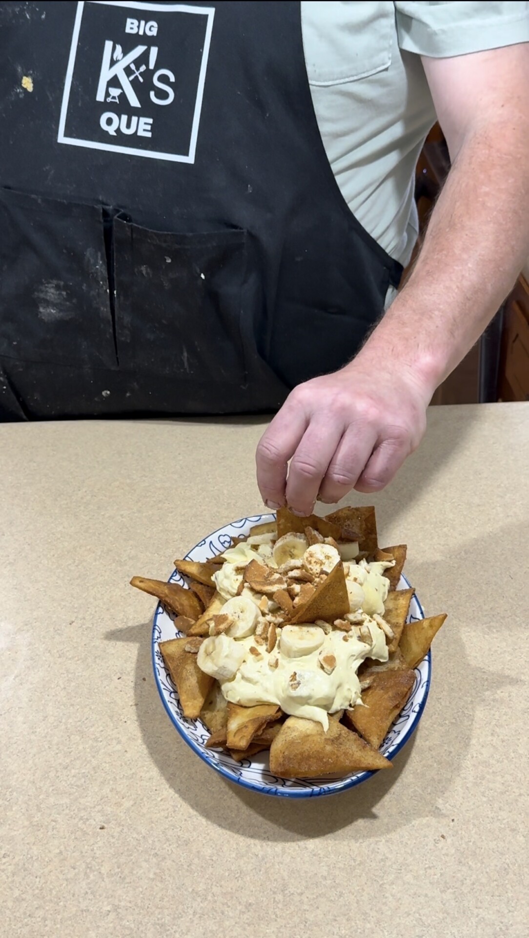 A person wearing a black apron with BIG KS QUE places chopped nuts on a bowl of fried pastry chips topped with banana slices and cream, set on a beige countertop.