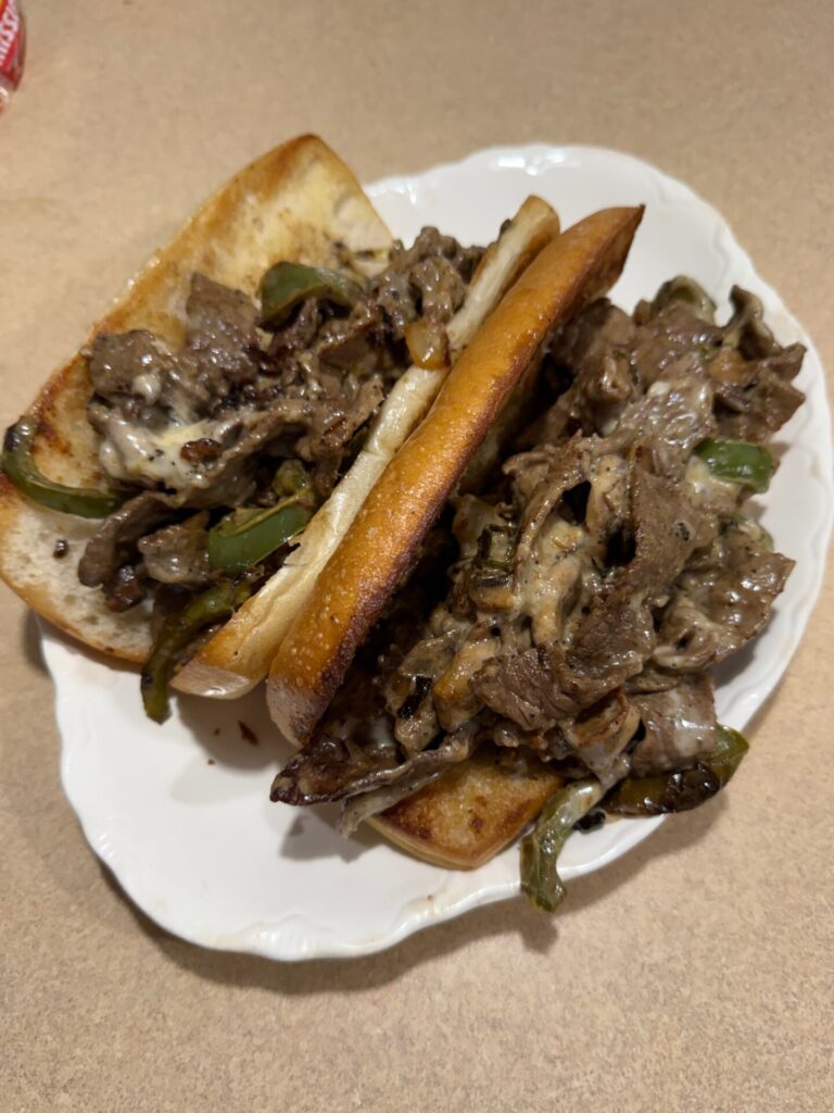 Two Philly cheesesteak sandwiches on a white plate, filled with sliced beef, melted cheese, green bell peppers, and onions, served on toasted hoagie rolls atop a beige countertop.