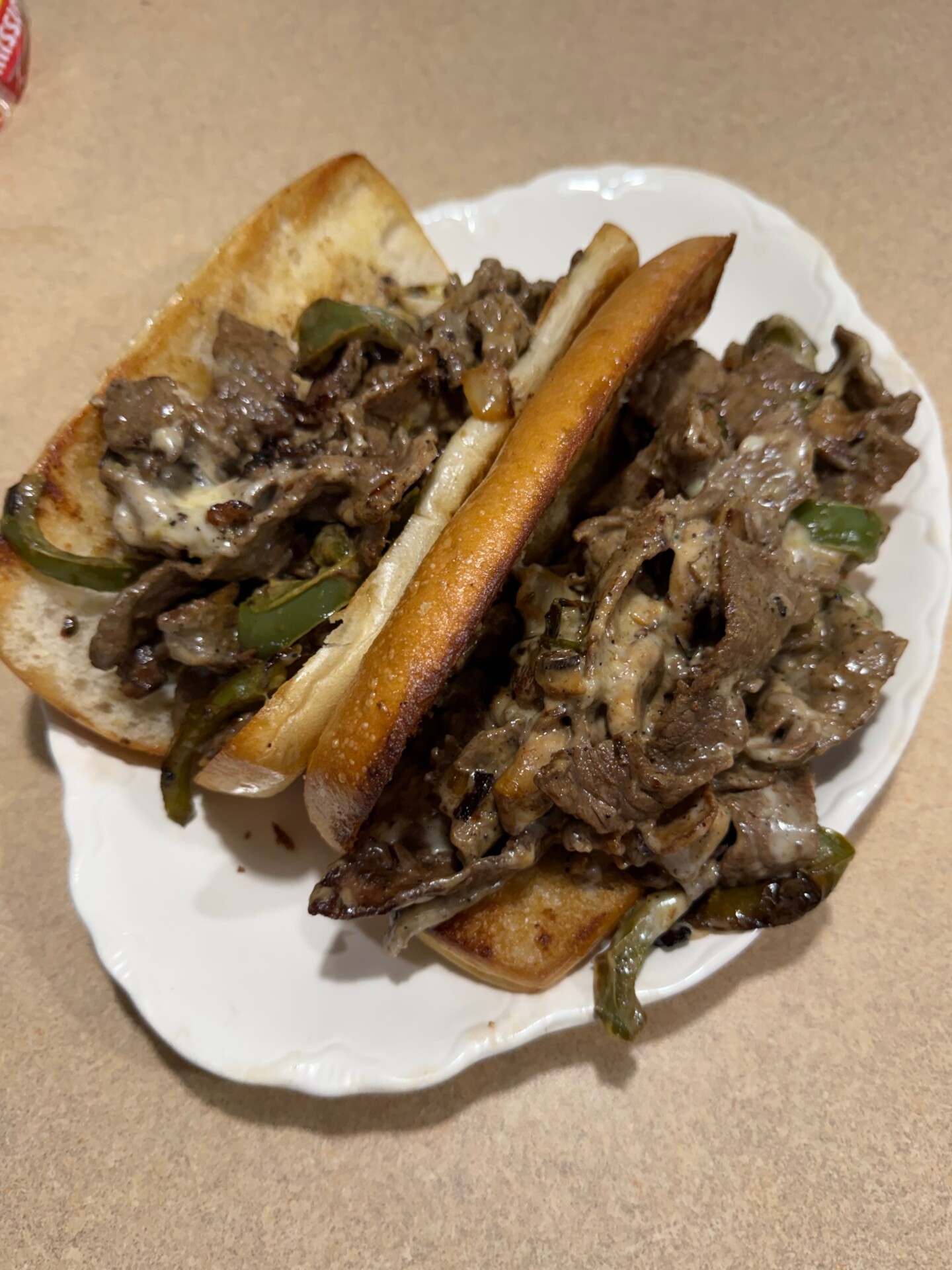 Two Philly cheesesteak sandwiches on a white plate, filled with sliced beef, melted cheese, green bell peppers, and onions, served on toasted hoagie rolls atop a beige countertop.