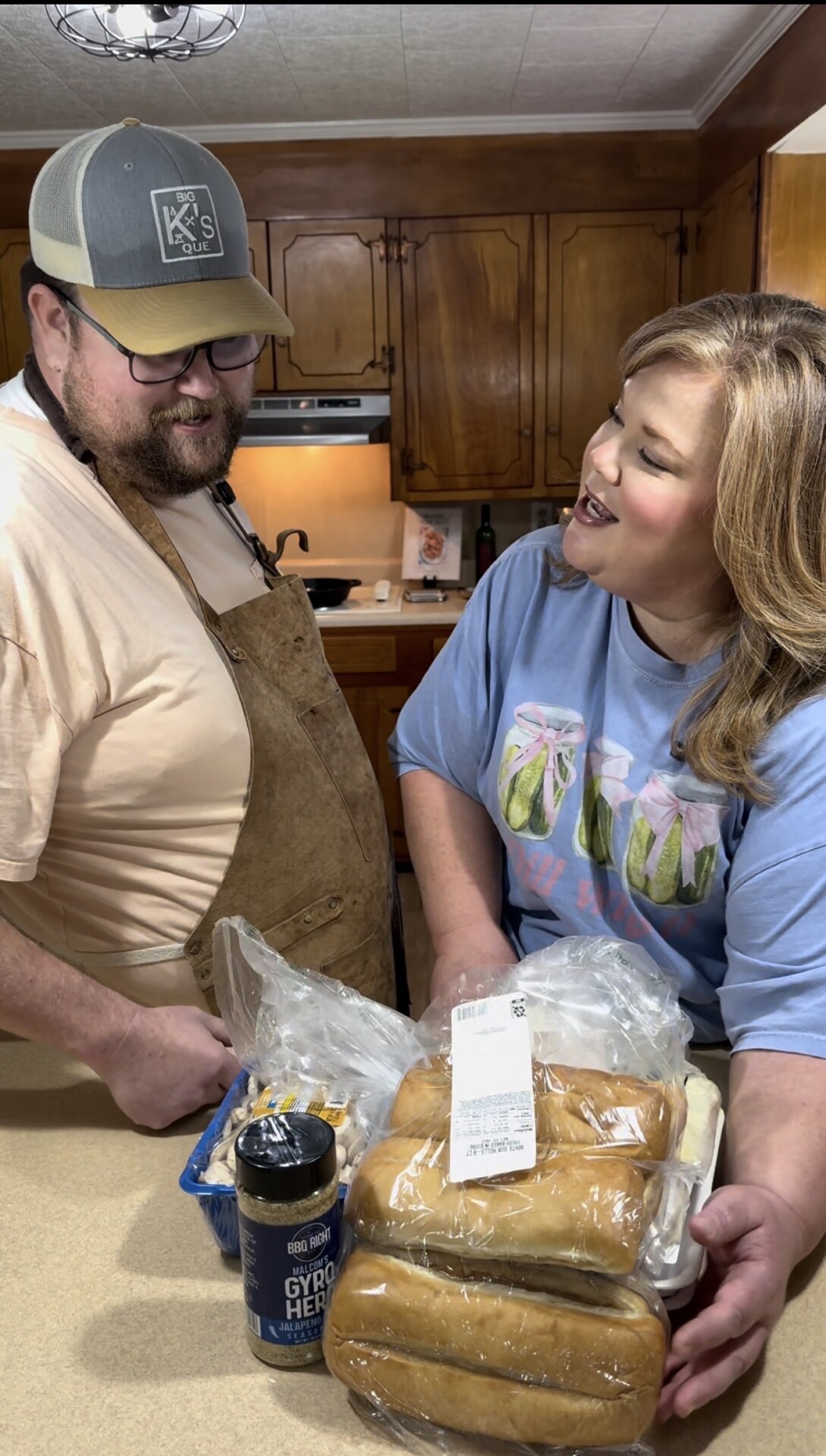 A man and woman stand together in a kitchen, smiling at each other. The counter in front of them holds a package of buns, a spice jar, and ingredients in plastic bags. The kitchen has wooden cabinets and warm lighting.