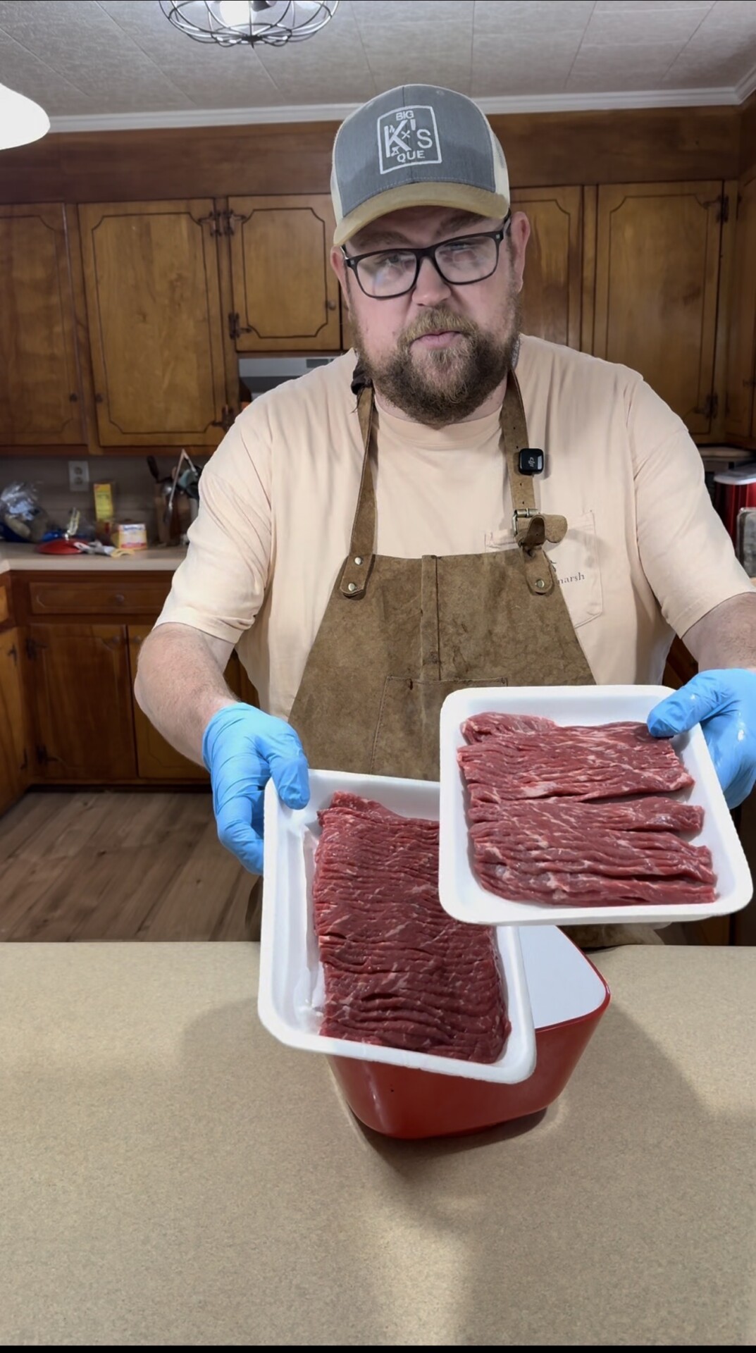 A man wearing a cap, glasses, and an apron stands in a kitchen, holding two trays of raw beef while wearing blue gloves. The background shows wooden cabinets and kitchen appliances.