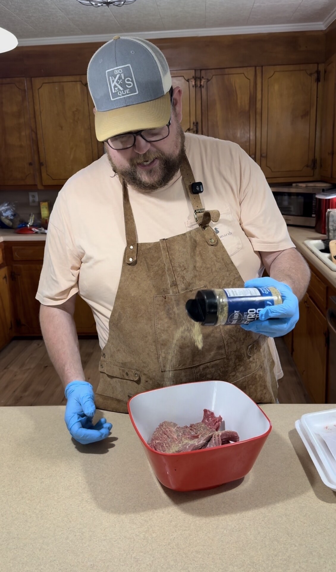 A bearded man wearing glasses, a cap, gloves, and an apron sprinkles seasoning onto raw meat in a red and white bowl on a kitchen counter. Wooden cabinets and kitchen appliances are visible in the background.