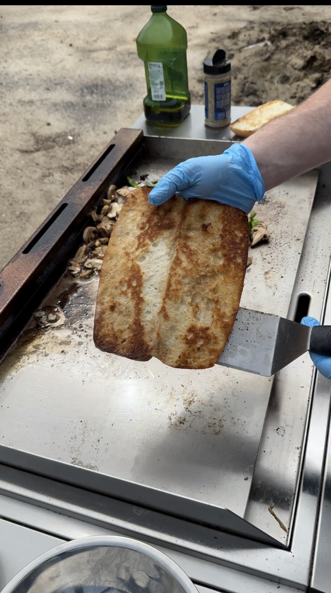 A person wearing blue gloves holds a toasted sandwich roll with a metal spatula over a flat-top grill, with mushrooms and other food items cooking nearby.