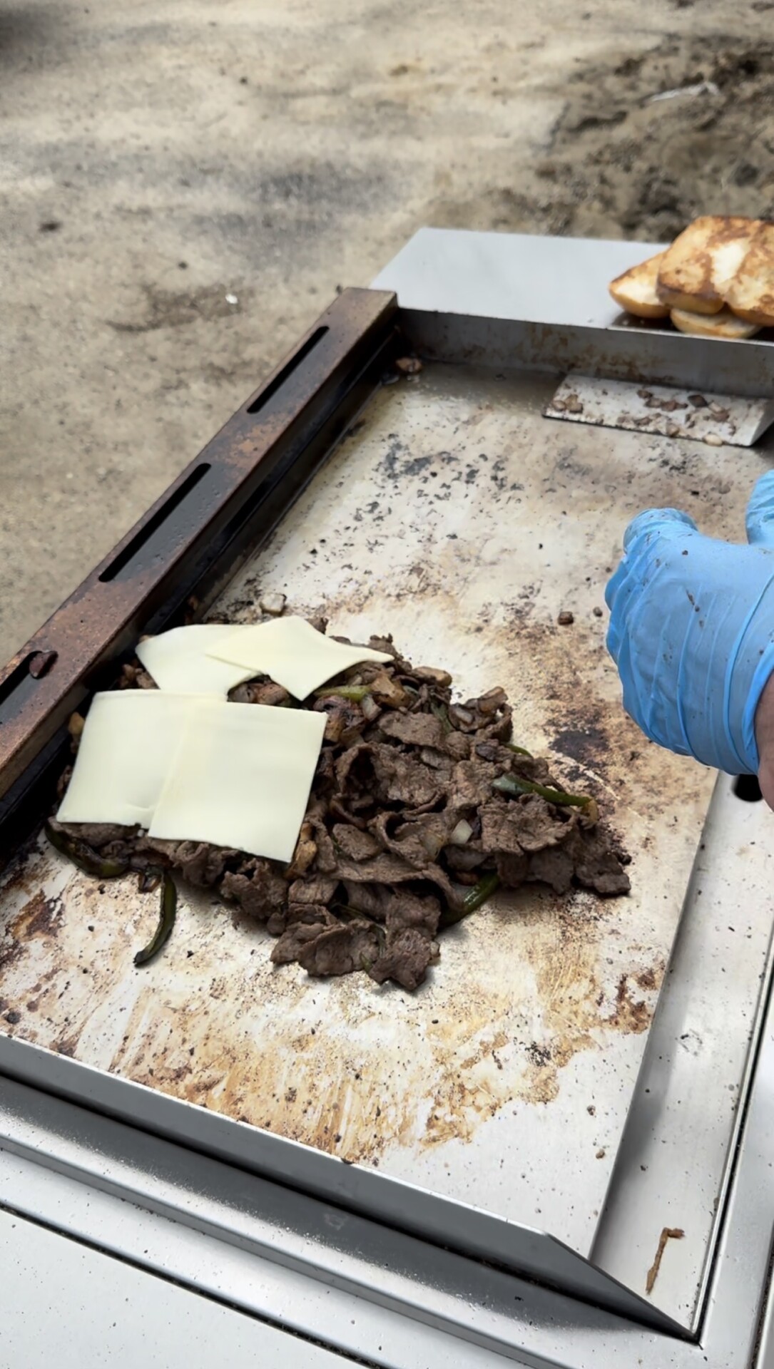 Slices of steak, onions, and green peppers cooking on a flat grill with pieces of white cheese on top. A person wearing blue gloves is working near the food, and toasted bread is visible in the background.