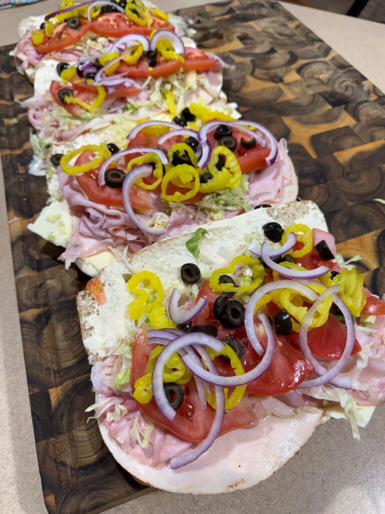 Four loaded sub sandwiches on a wooden board, topped with sliced tomatoes, red onions, black olives, banana peppers, shredded lettuce, ham, turkey, and cheese.