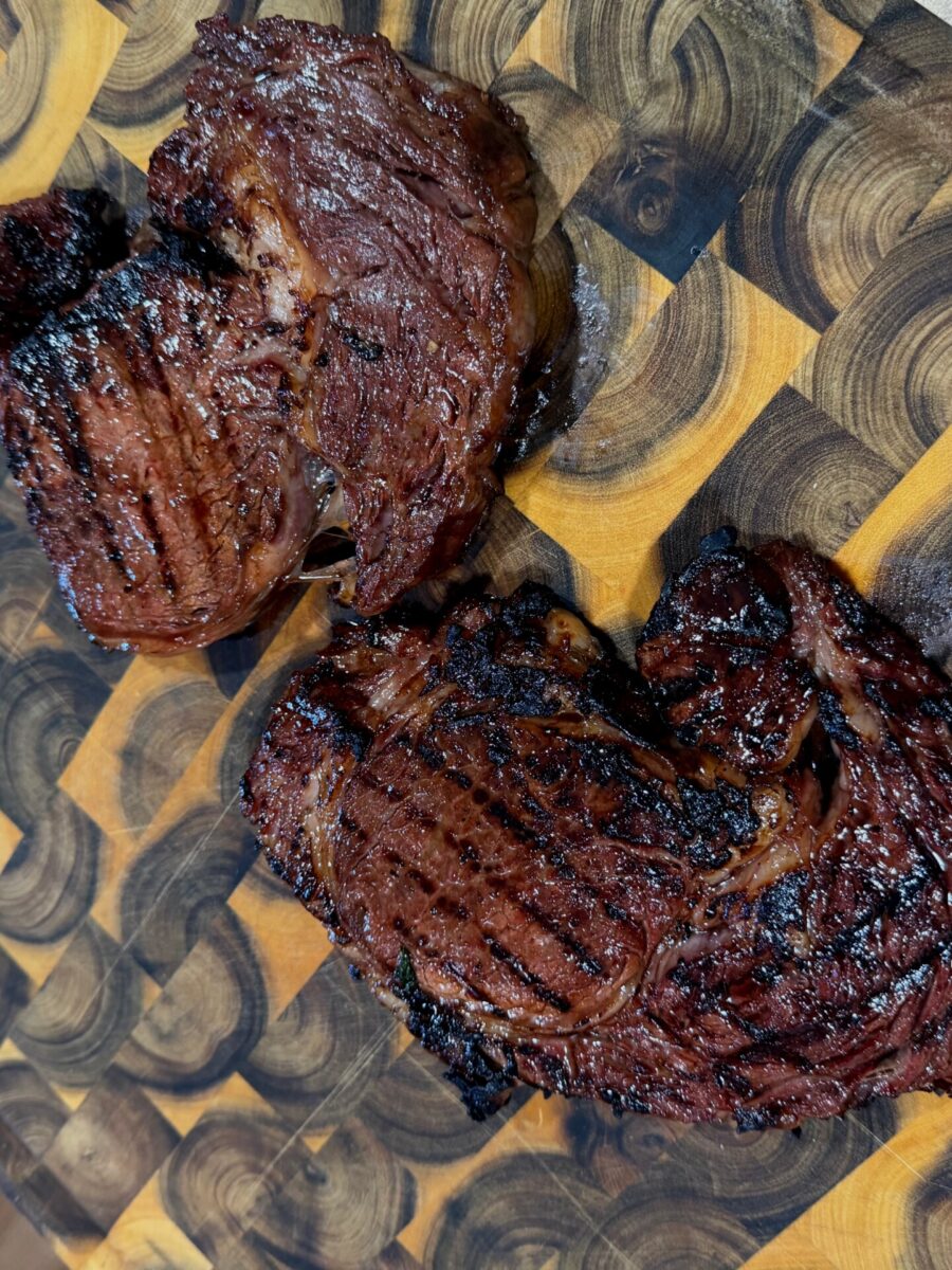 Two grilled ribeye steaks with dark grill marks rest on a wooden cutting board with a circular pattern. The steaks appear juicy and well-cooked with a charred, caramelized exterior.