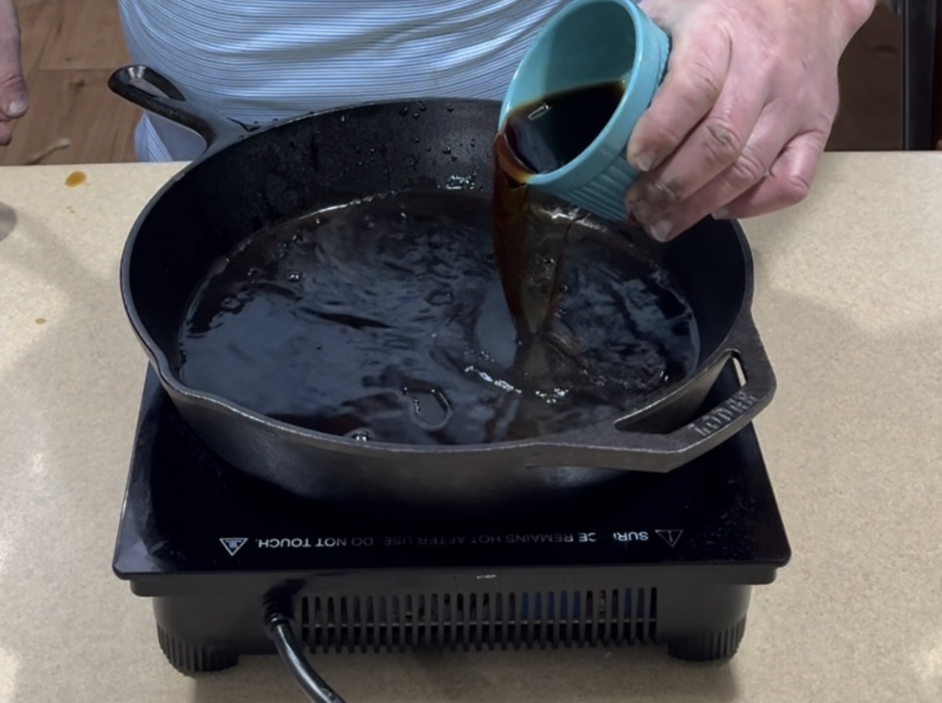 A person pours a dark liquid from a small blue cup into a cast-iron skillet on an electric cooktop.