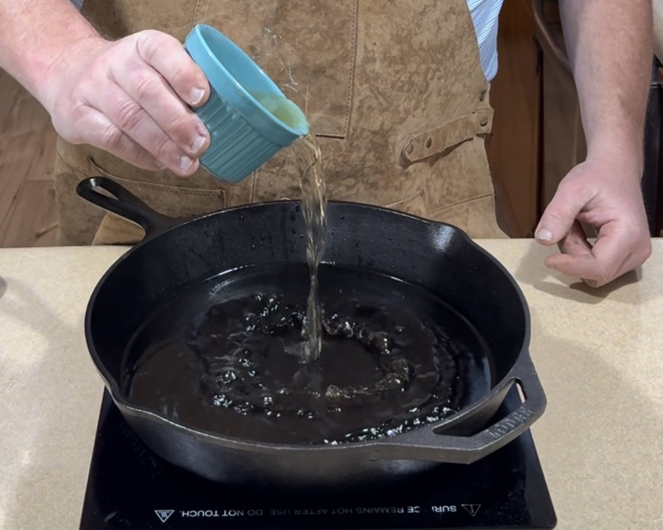 A person in a brown apron pours liquid from a blue cup into a black cast iron skillet on a stovetop.