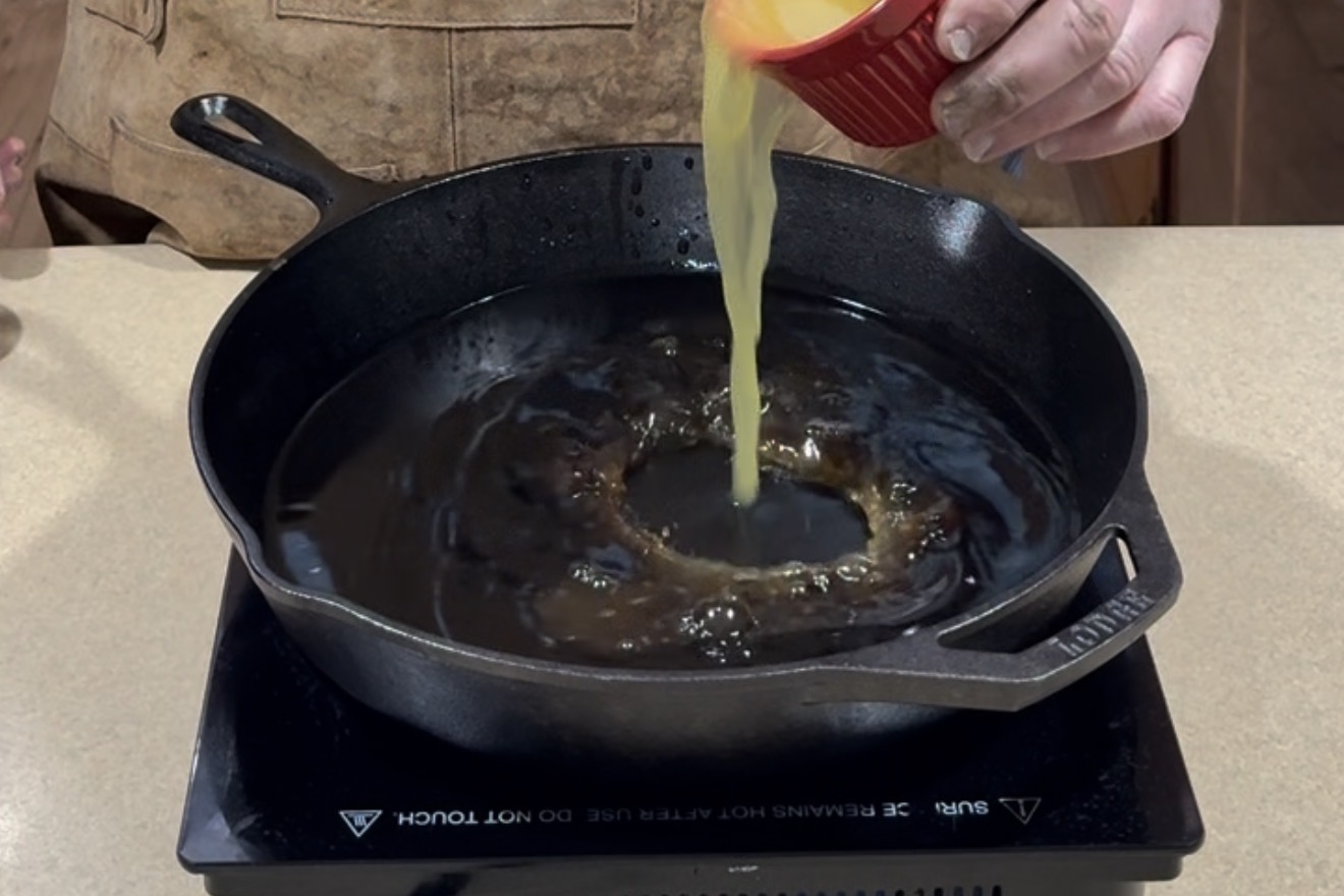 A person pours beaten eggs from a red ramekin into a hot, greased cast iron skillet on a stovetop.