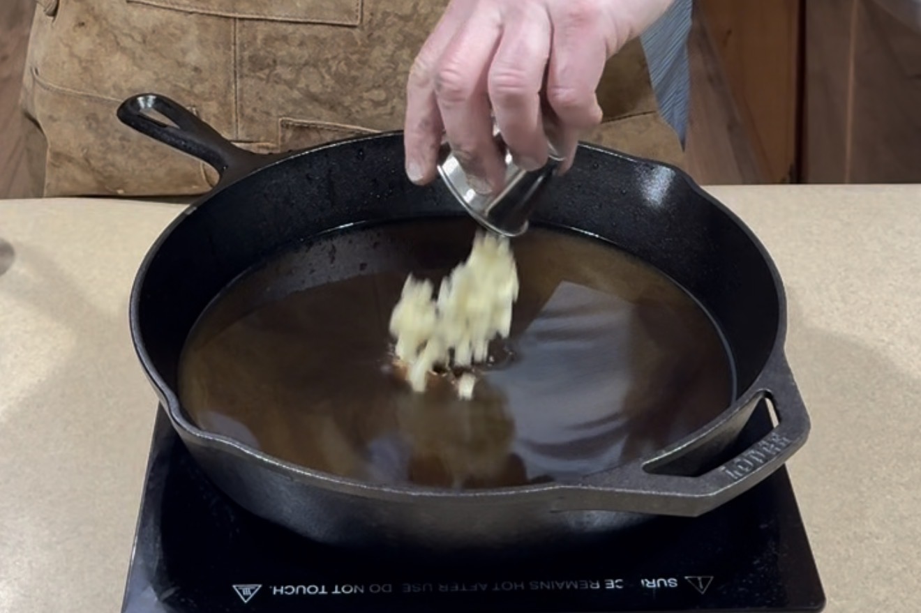 A person adds chopped garlic from a small container into a black cast iron skillet filled with oil, which is placed on an electric stovetop.