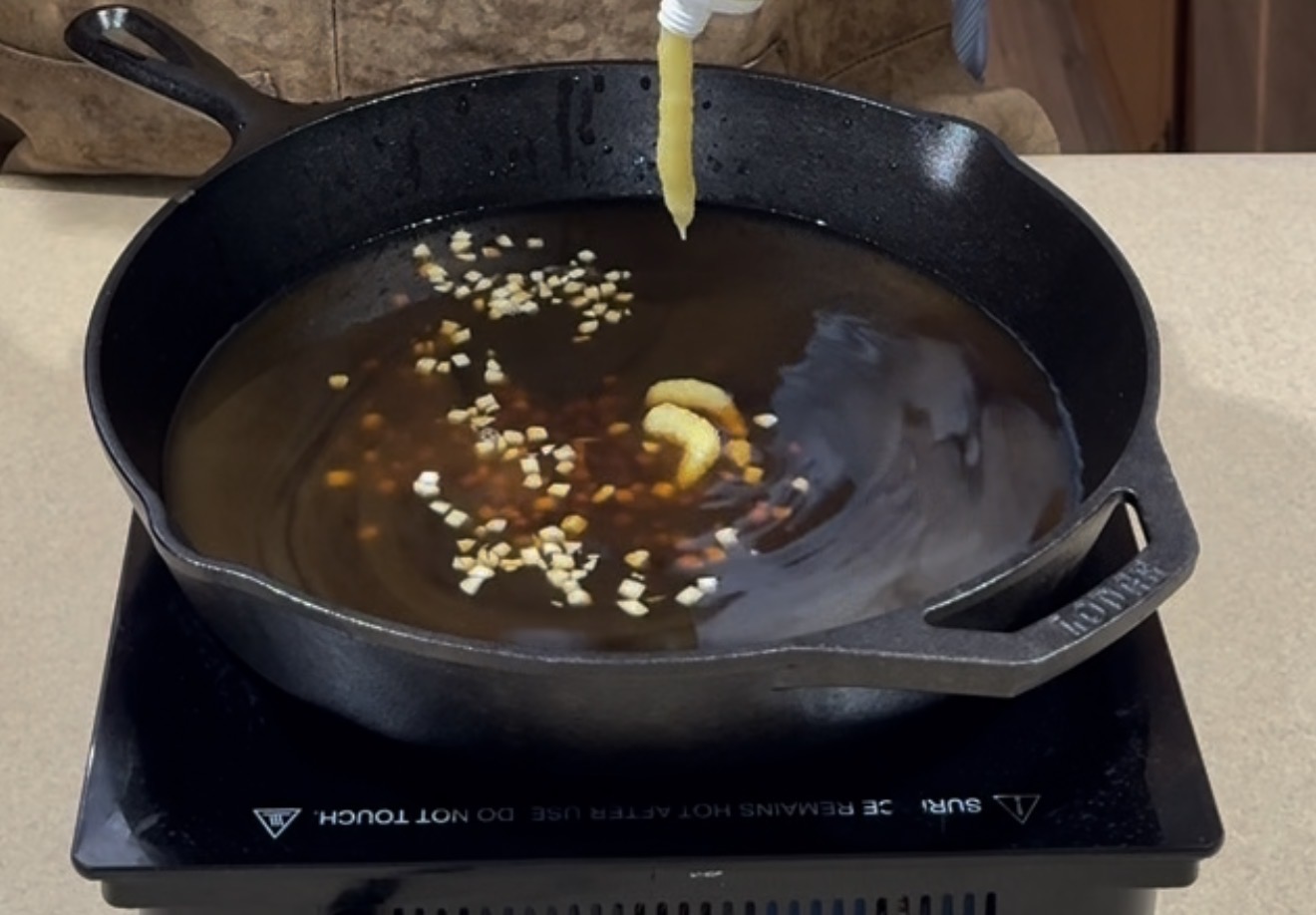 A cast iron skillet on an induction cooktop contains oil, minced garlic, and sauce, with paste being squeezed into the pan from above.